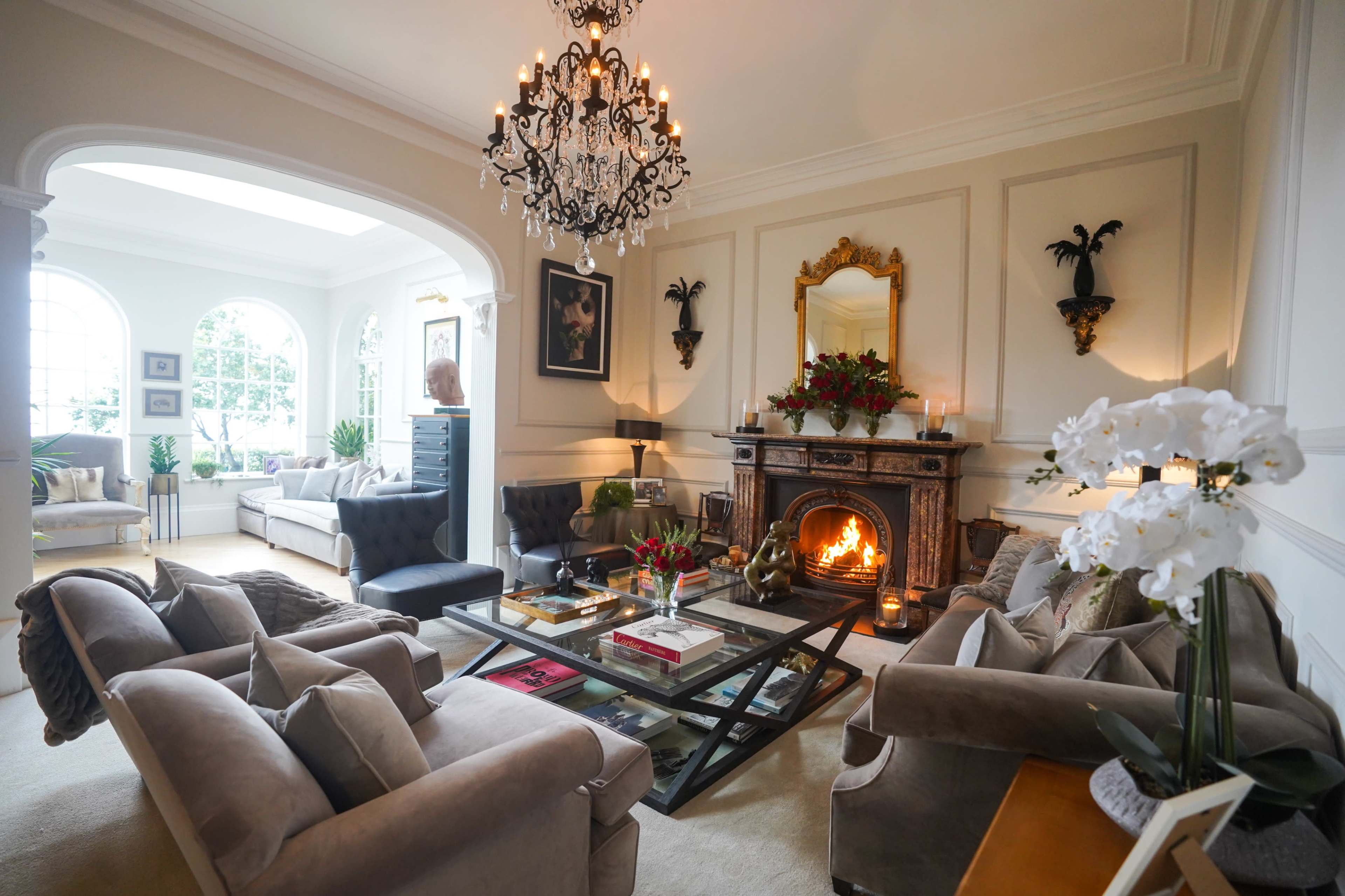 A stylish living room features a chandelier, a fireplace with an ornate mantel, and comfortable seating arranged around a central glass coffee table.