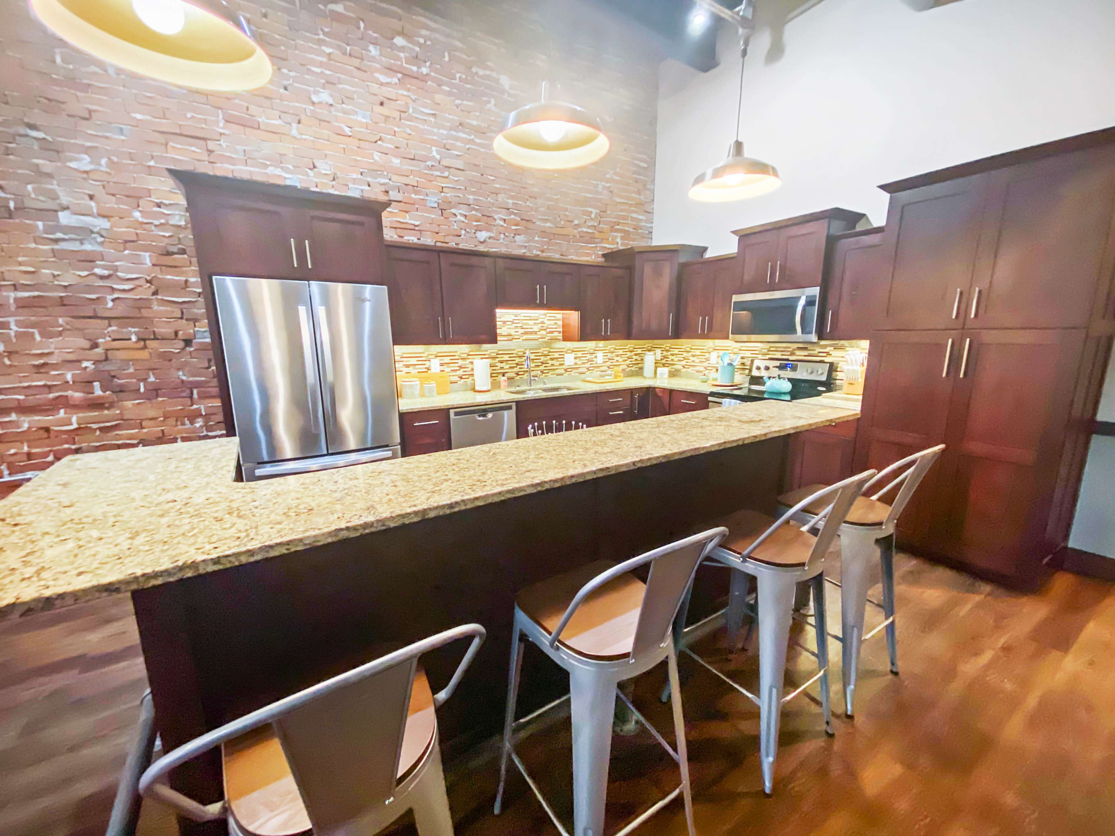 A modern kitchen features dark wood cabinets, a large granite island with seating, and exposed brick walls.