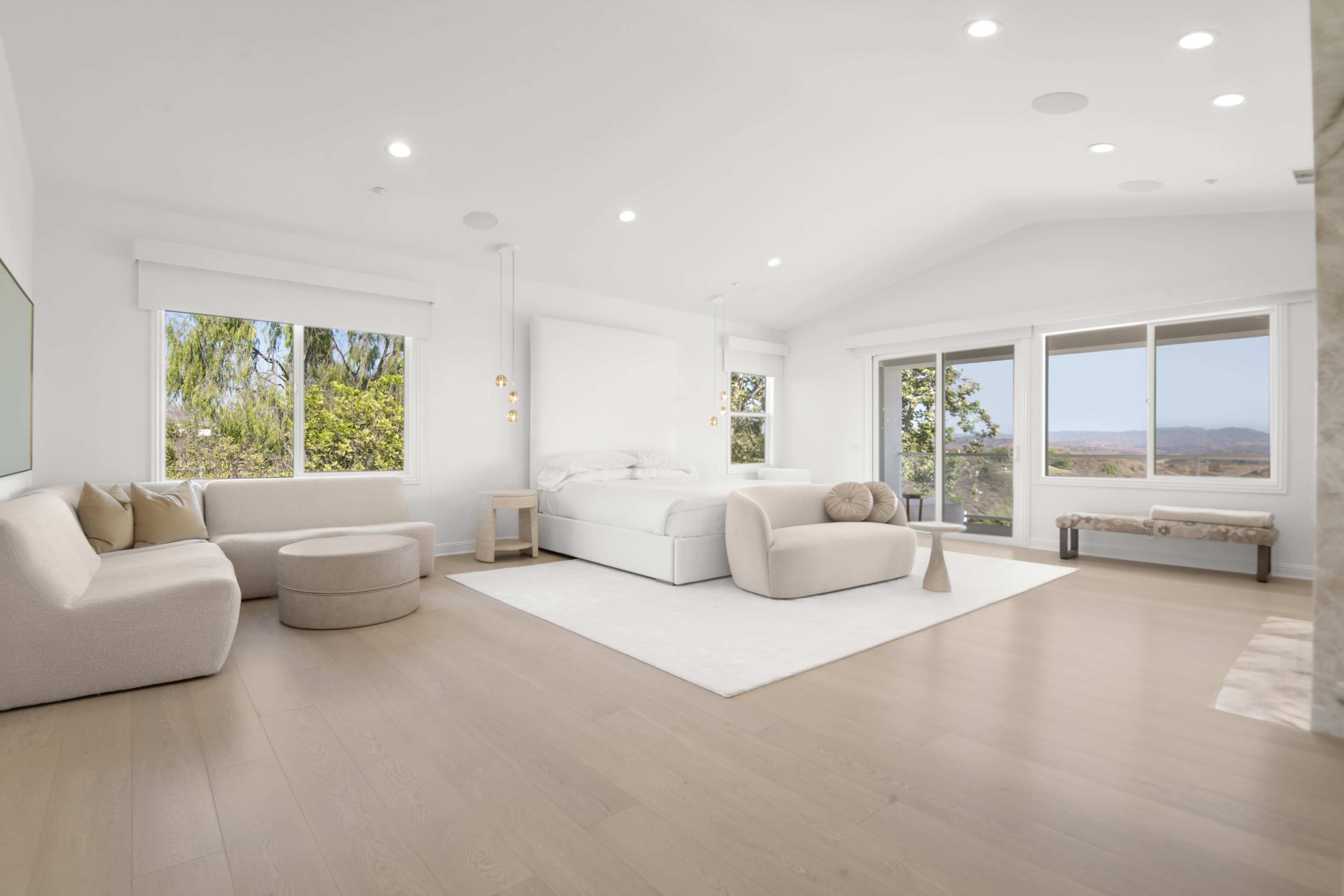 A spacious, bright room features a bed, a circular chair, and a small side table, with large windows overlooking a landscape.