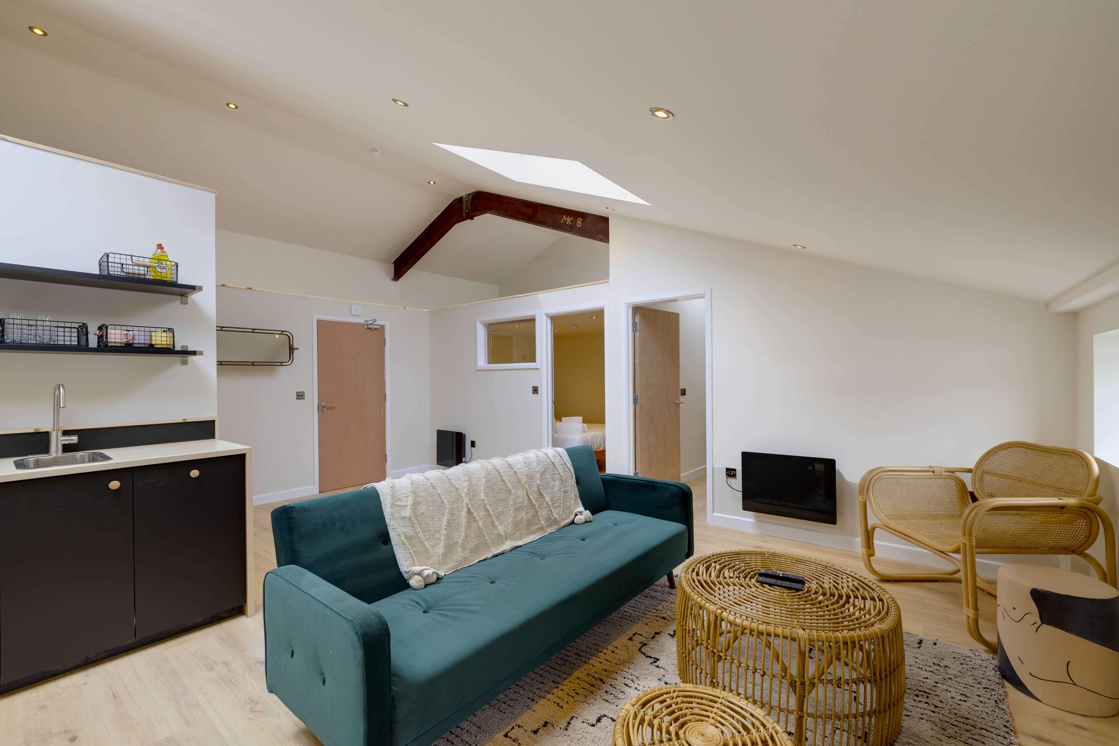 A modern living space features a teal sofa, a round rattan coffee table, and a wall-mounted TV, with natural light streaming in through a skylight.
