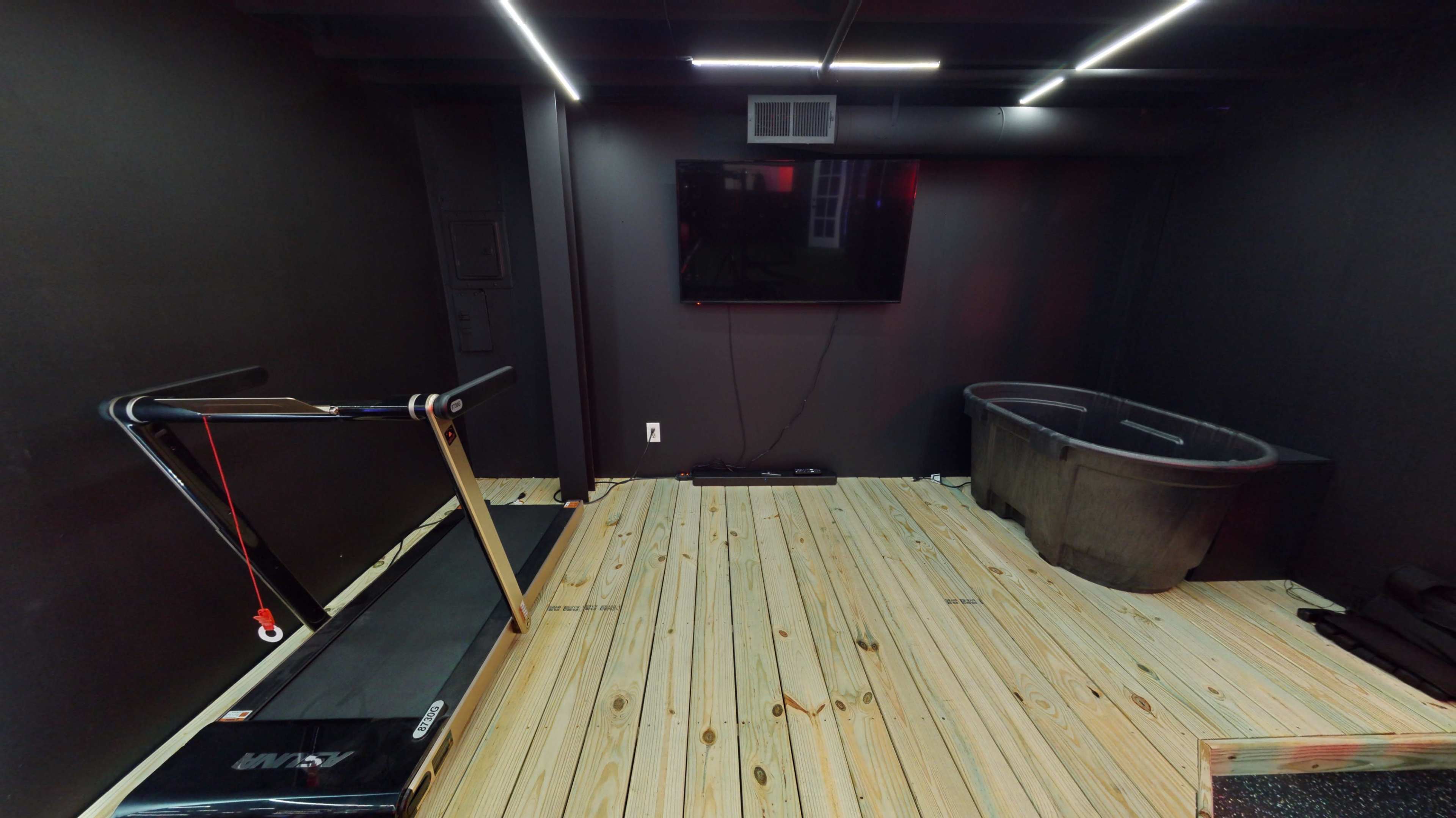 A fitness room with a treadmill, a large black tub, and a wall-mounted television set against dark walls and wooden flooring.