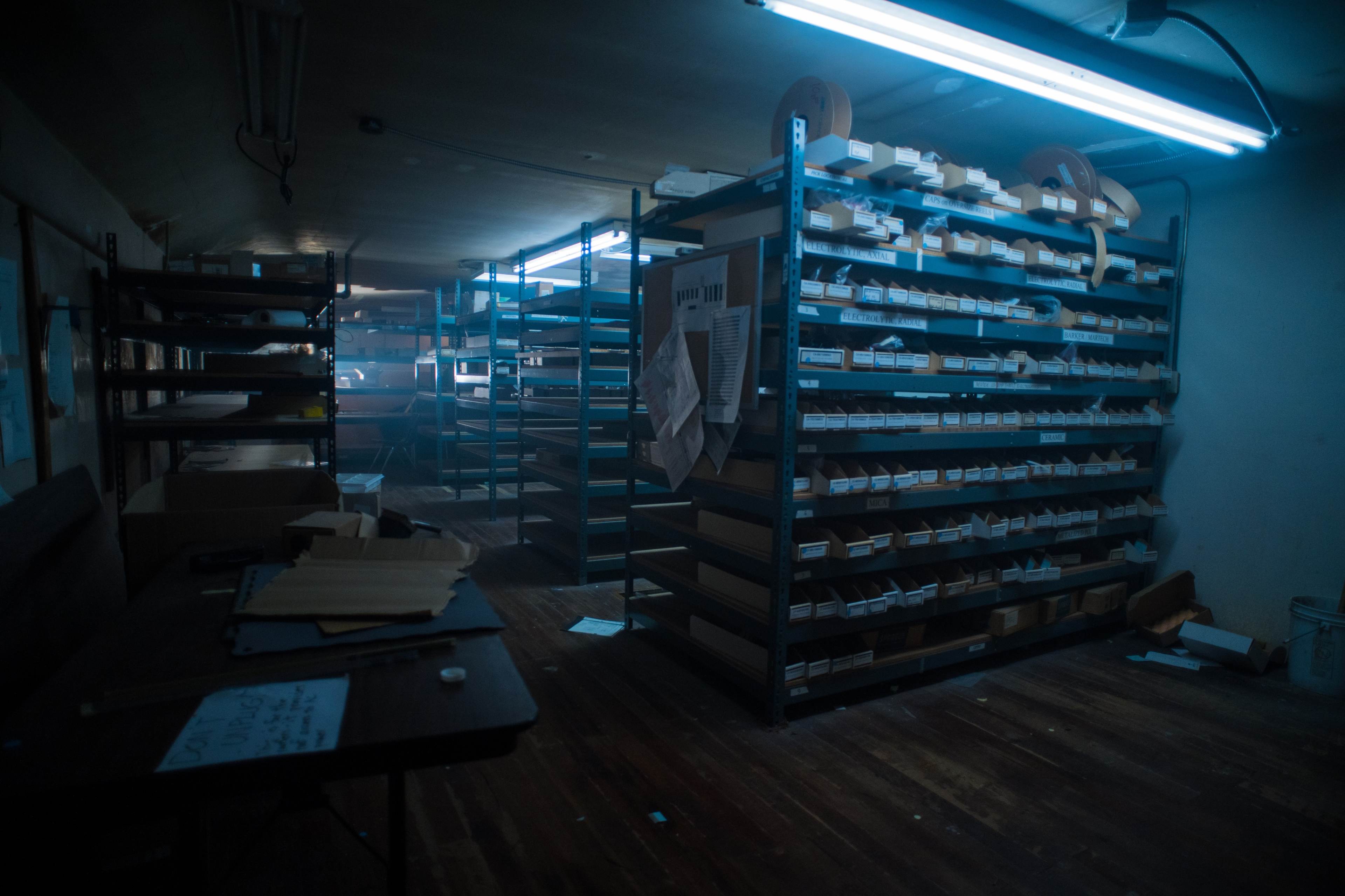 A dimly lit storage room filled with metal shelving units stacked with various labeled boxes and scattered papers on a wooden floor.