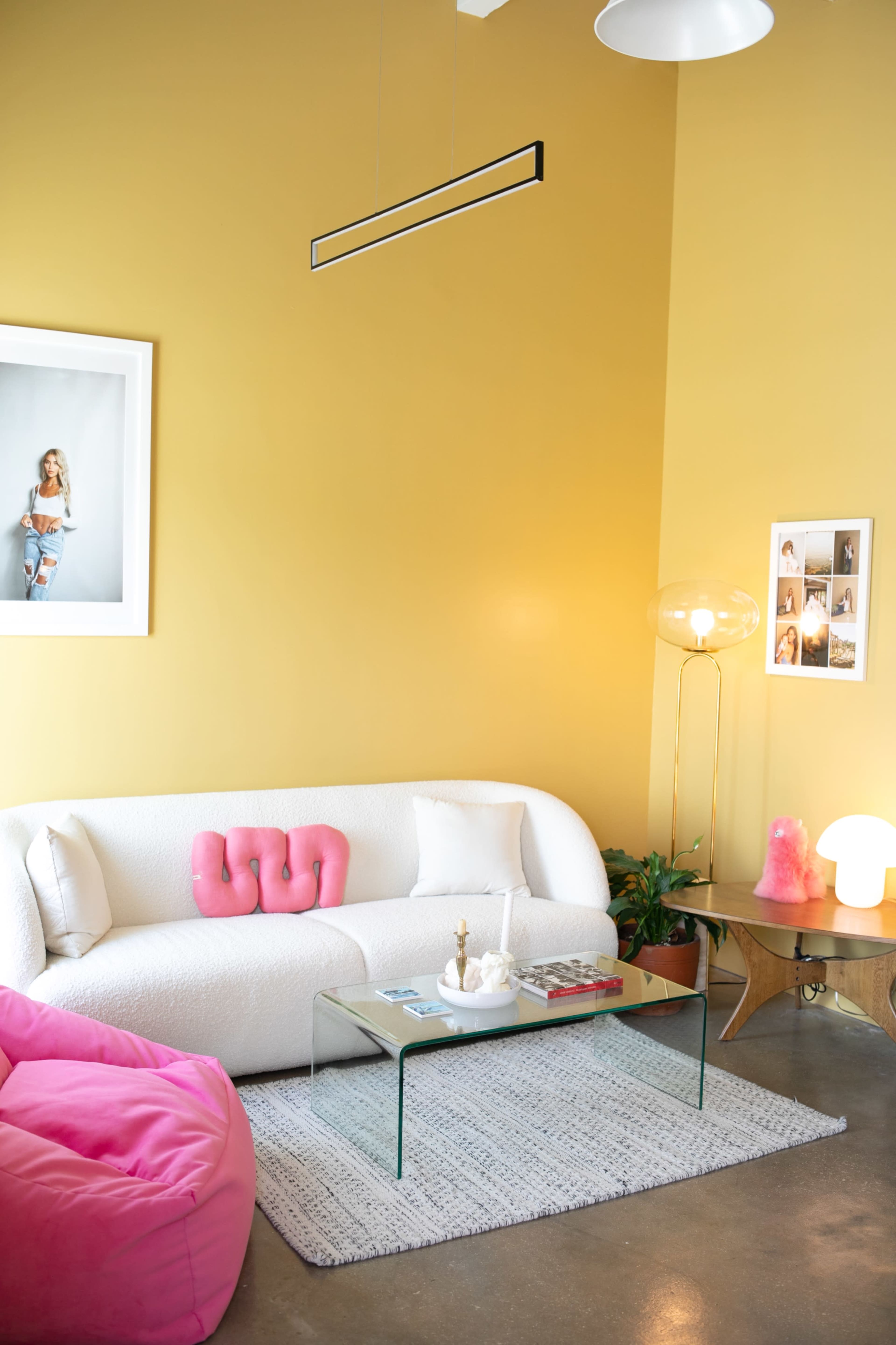 The image shows a cozy living room with a white sofa, a glass coffee table, and pink accents, set against a yellow wall and featuring a mix of decorative lighting.