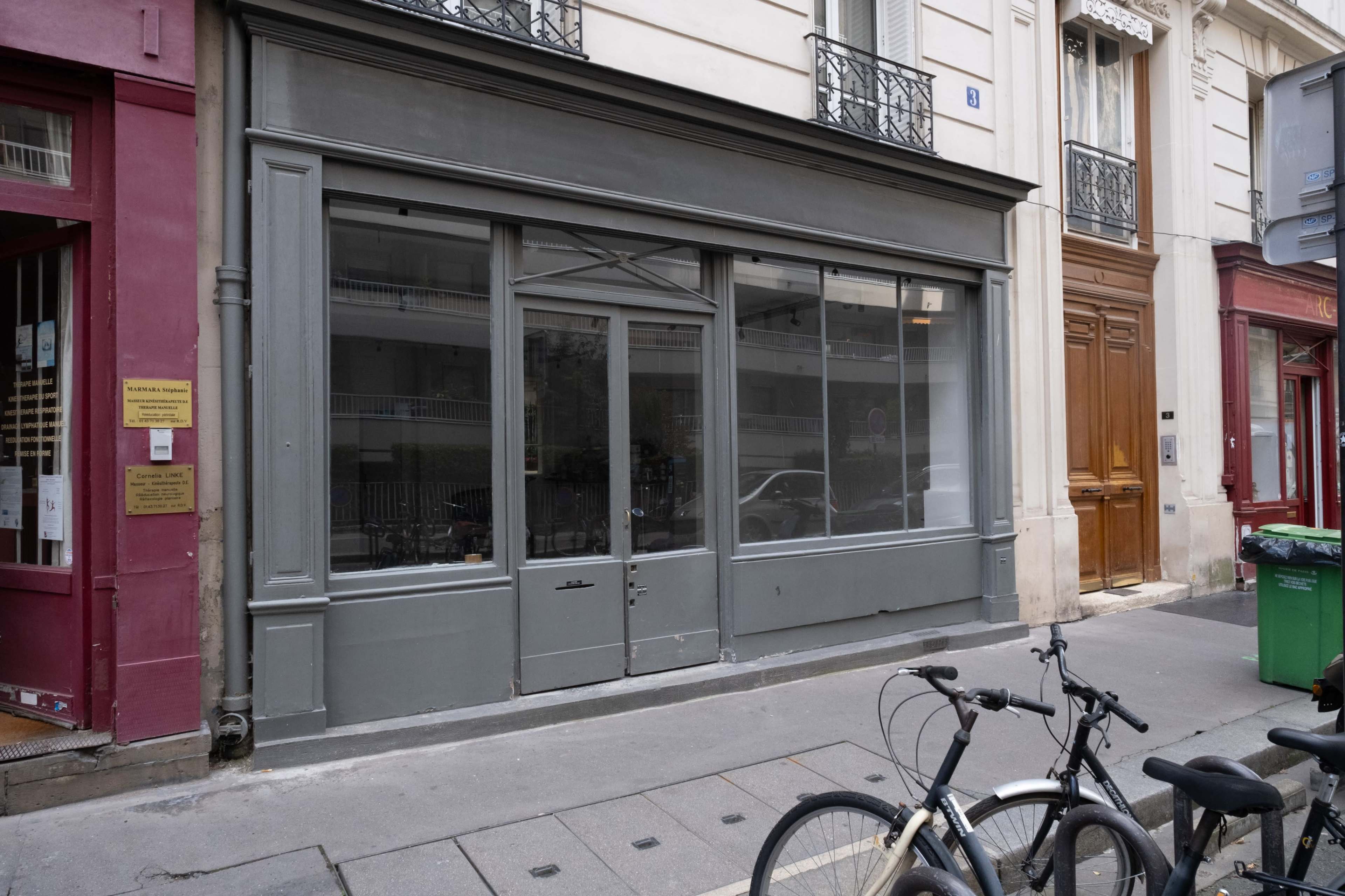The image shows a vacant storefront with a gray facade and large windows, located on a city street alongside parked bicycles.