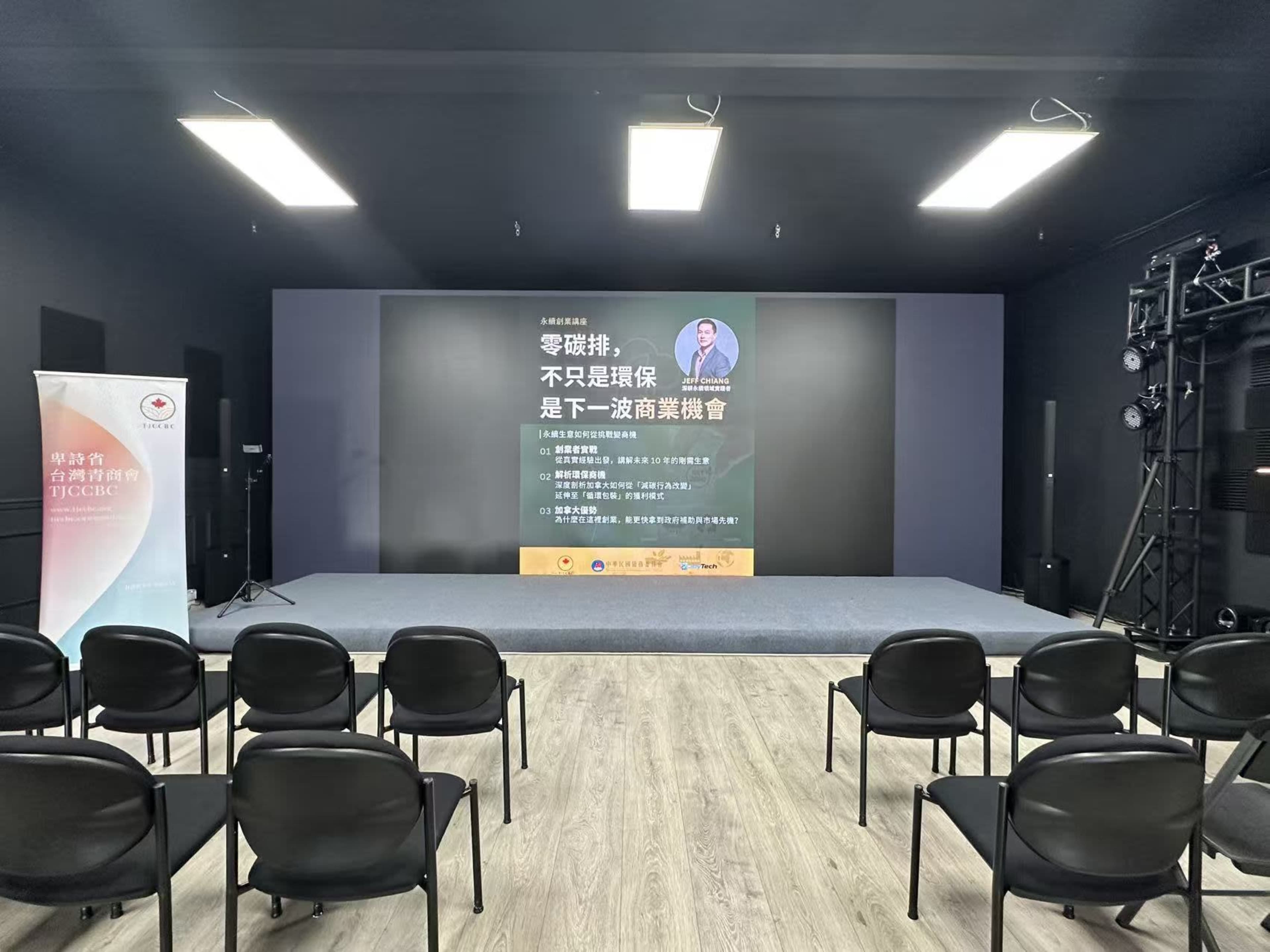 The image shows a blank stage set up for an event, featuring a large backdrop with text and a seating area filled with black chairs in front of it.