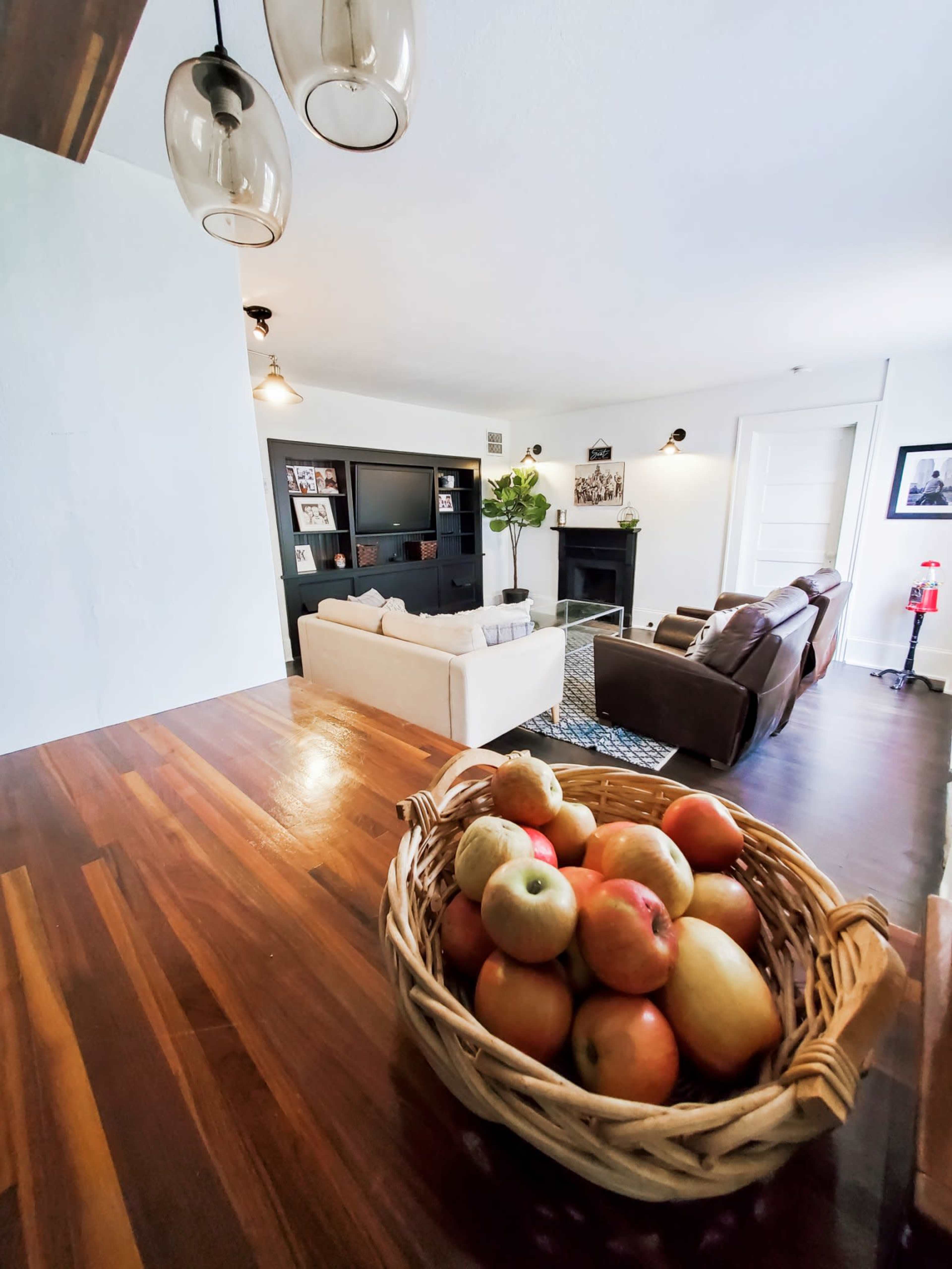 A bowl of apples rests on a wooden table in the foreground, while a living room with a white sofa and dark leather chairs is visible in the background.