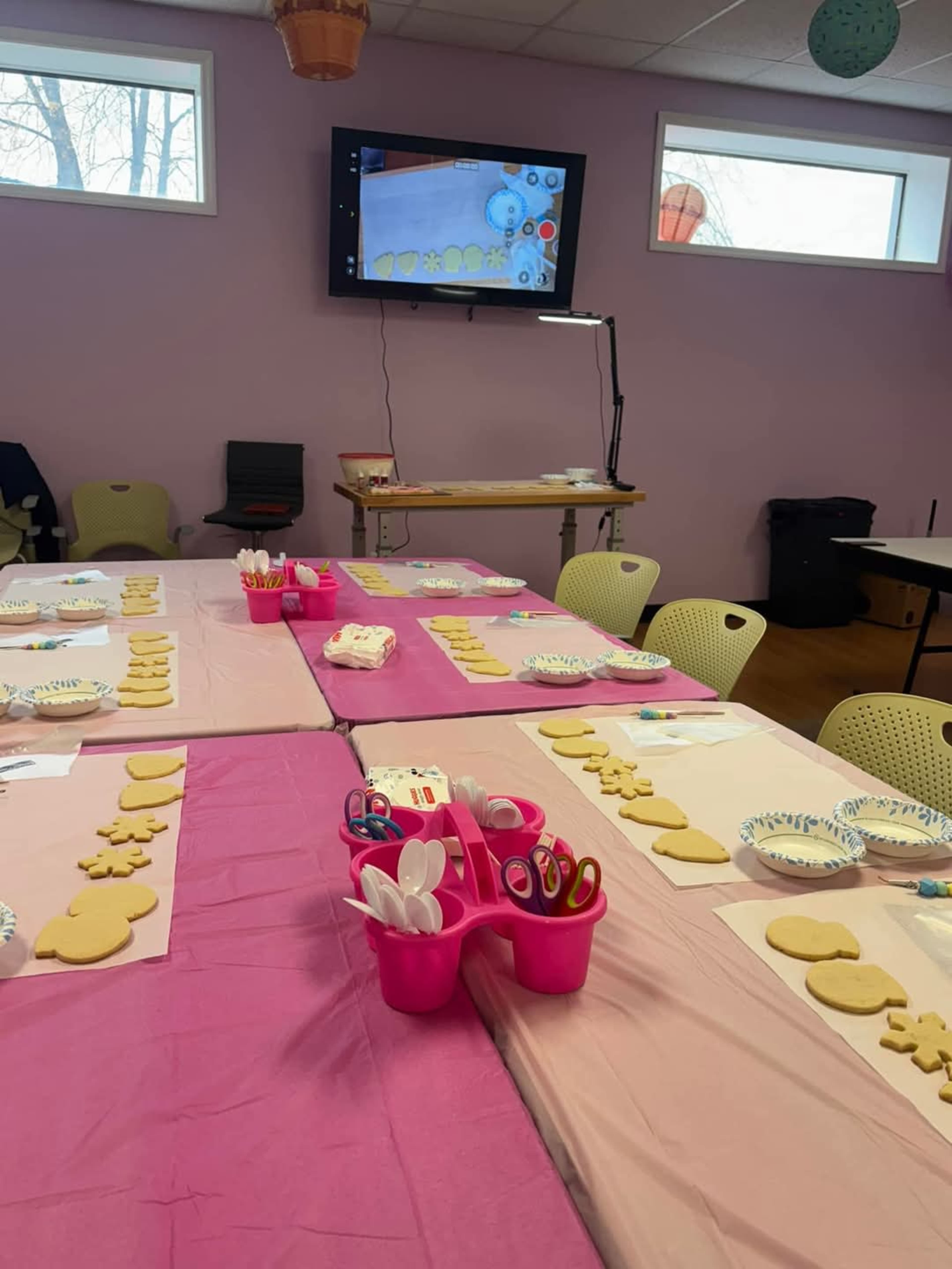 A brightly decorated room features tables set with cookie-cutting supplies, including dough shapes and colorful utensils, alongside a monitor displaying instructions.