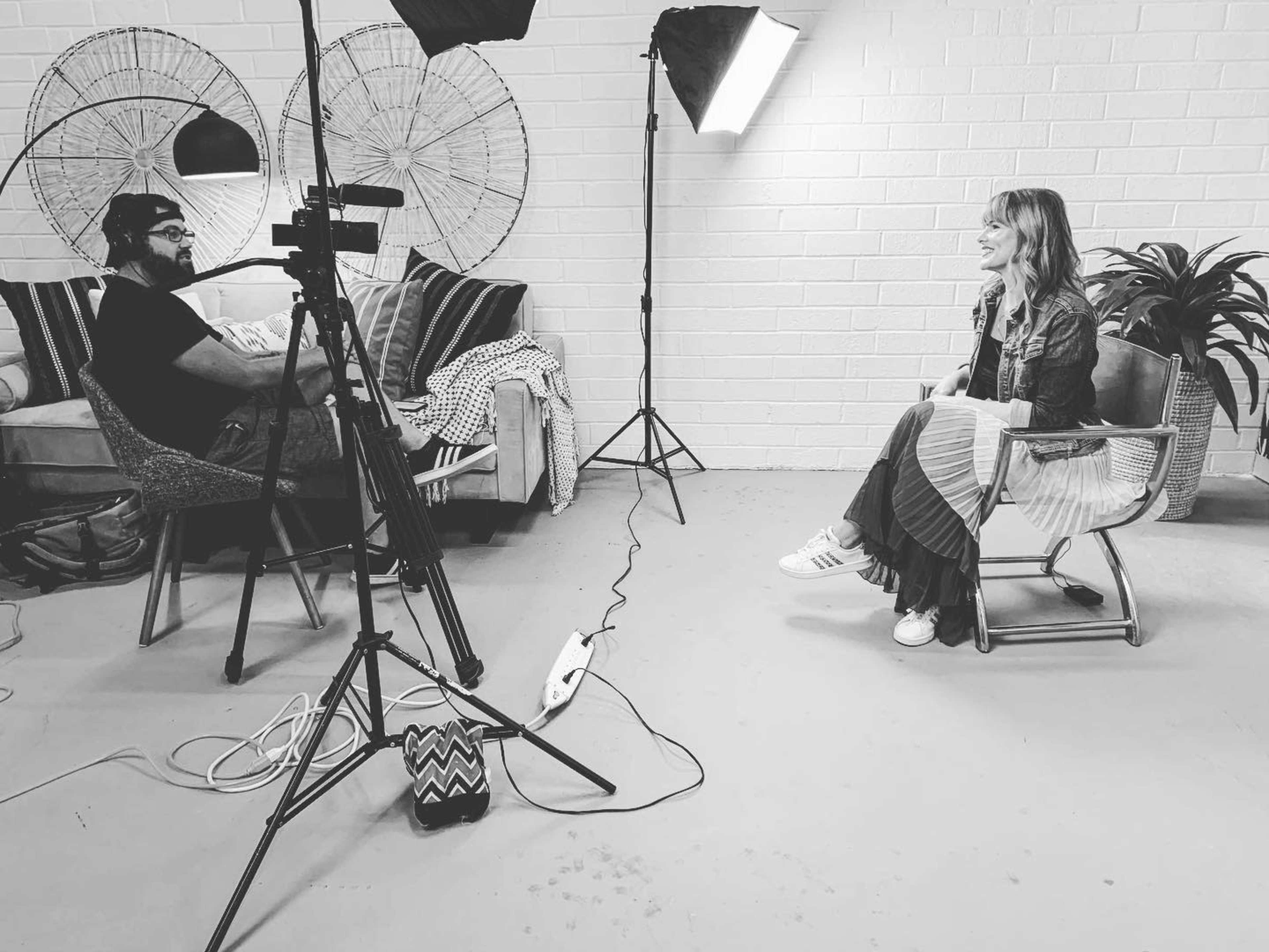 A person sits in a chair during an interview while another person operates a camera on a tripod in a well-lit room with a couch and decorative items.