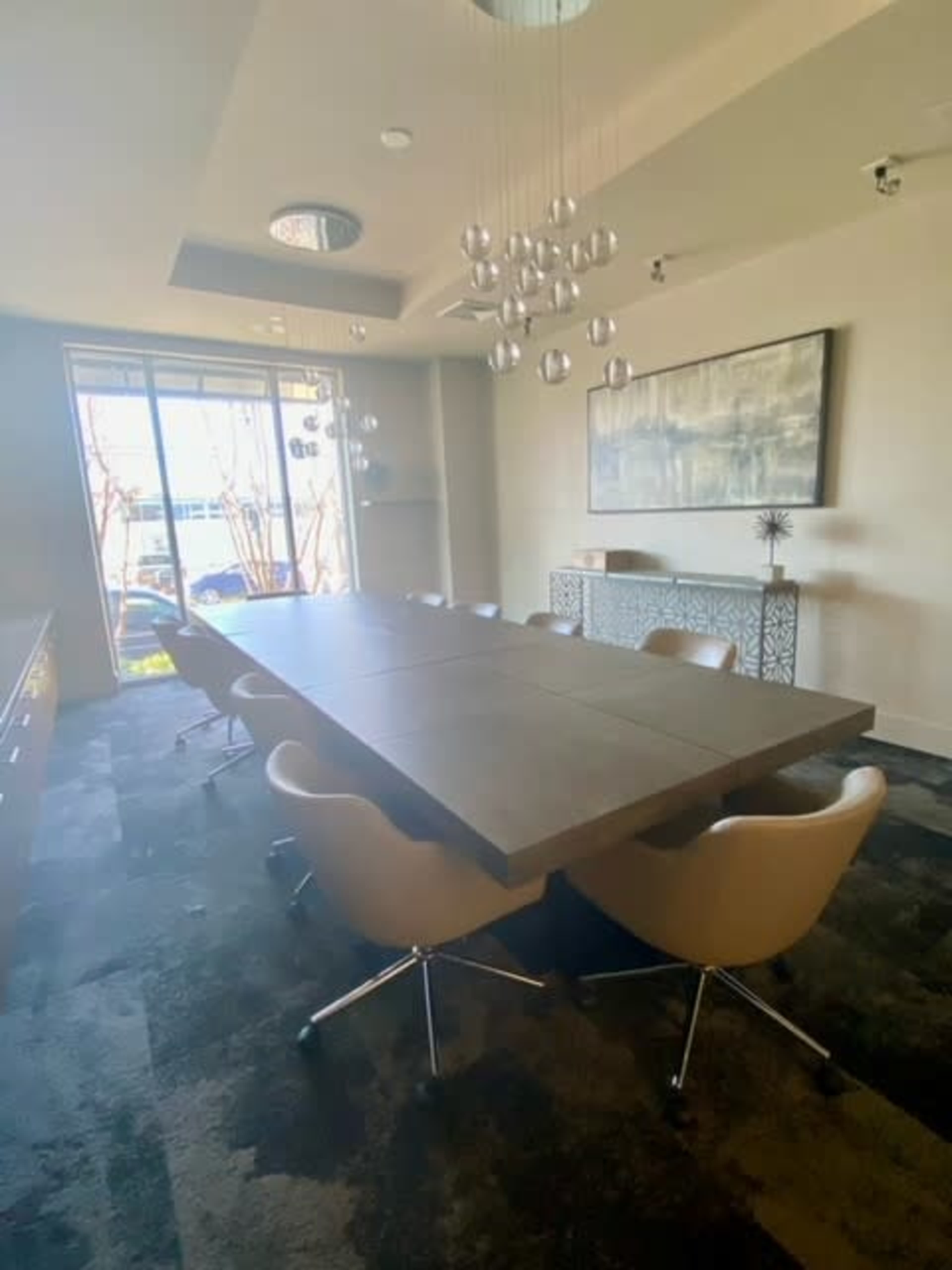 A modern conference room features a large rectangular table surrounded by chairs, with a window providing natural light and decorative pendant lights overhead.
