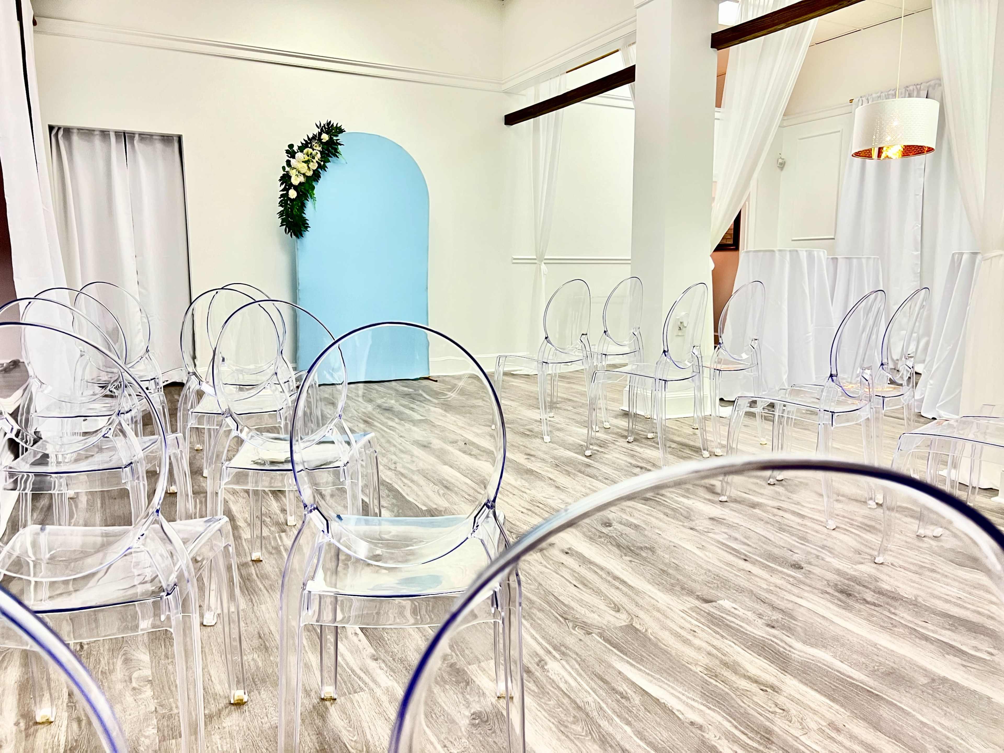 A spacious room is set up with clear acrylic chairs arranged in rows facing a blue arch and decorative greenery on one side.