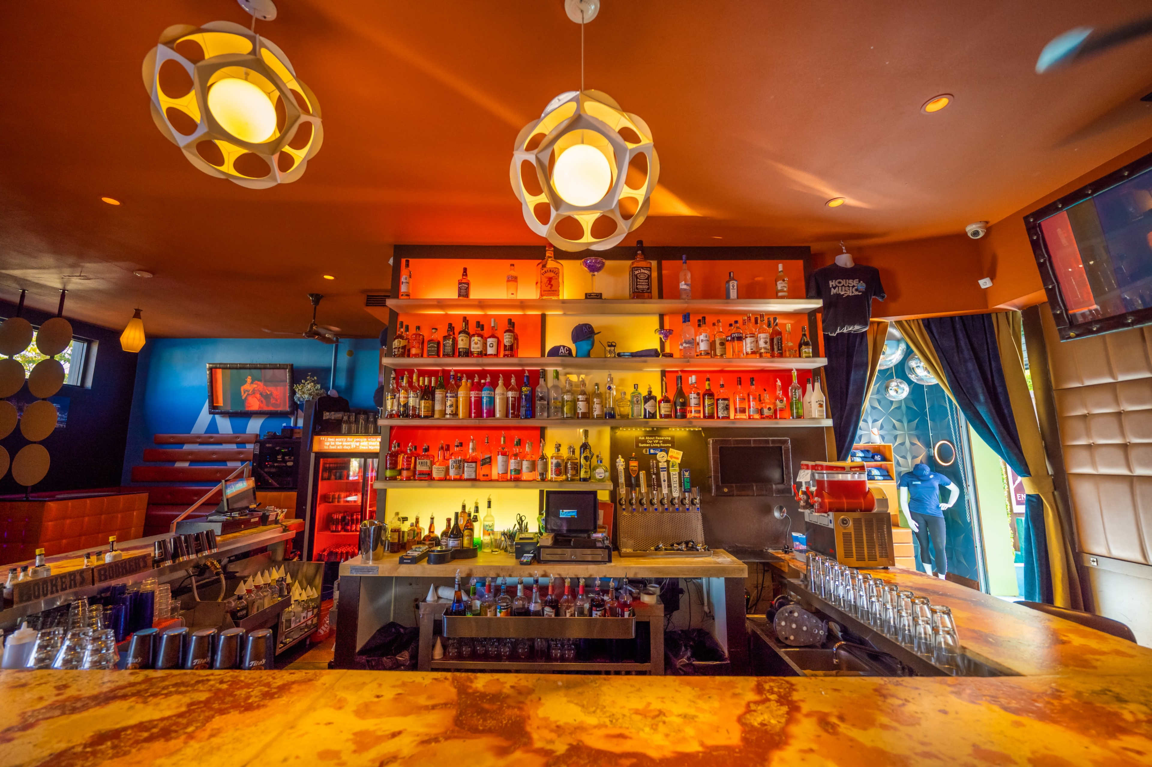 A well-stocked bar displays various bottles of liquor on brightly lit shelves with modern pendant lighting overhead.