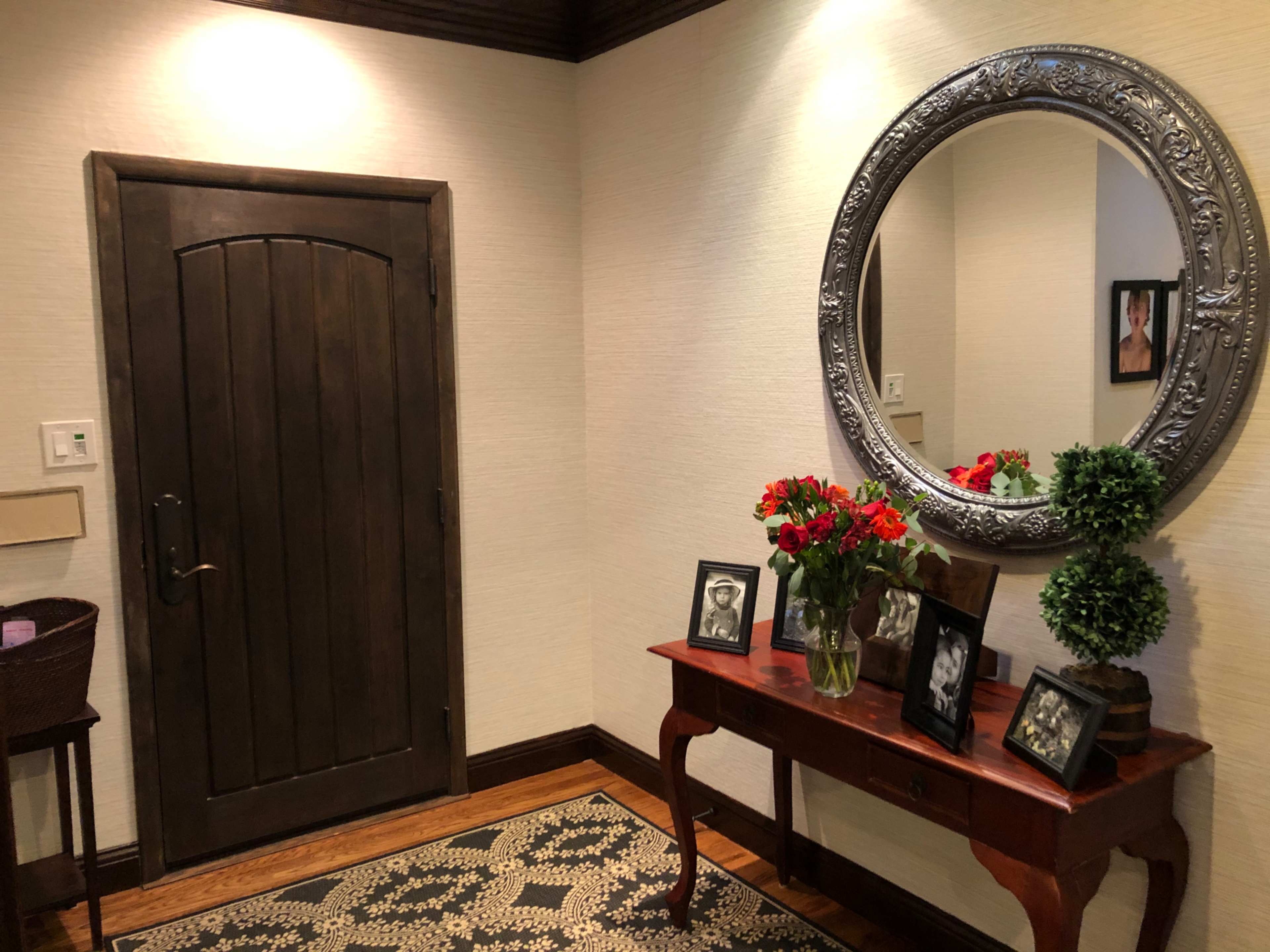 A hallway entrance featuring a round silver mirror above a wooden console table adorned with framed photos and a vase of flowers, with a dark wooden door to the left.