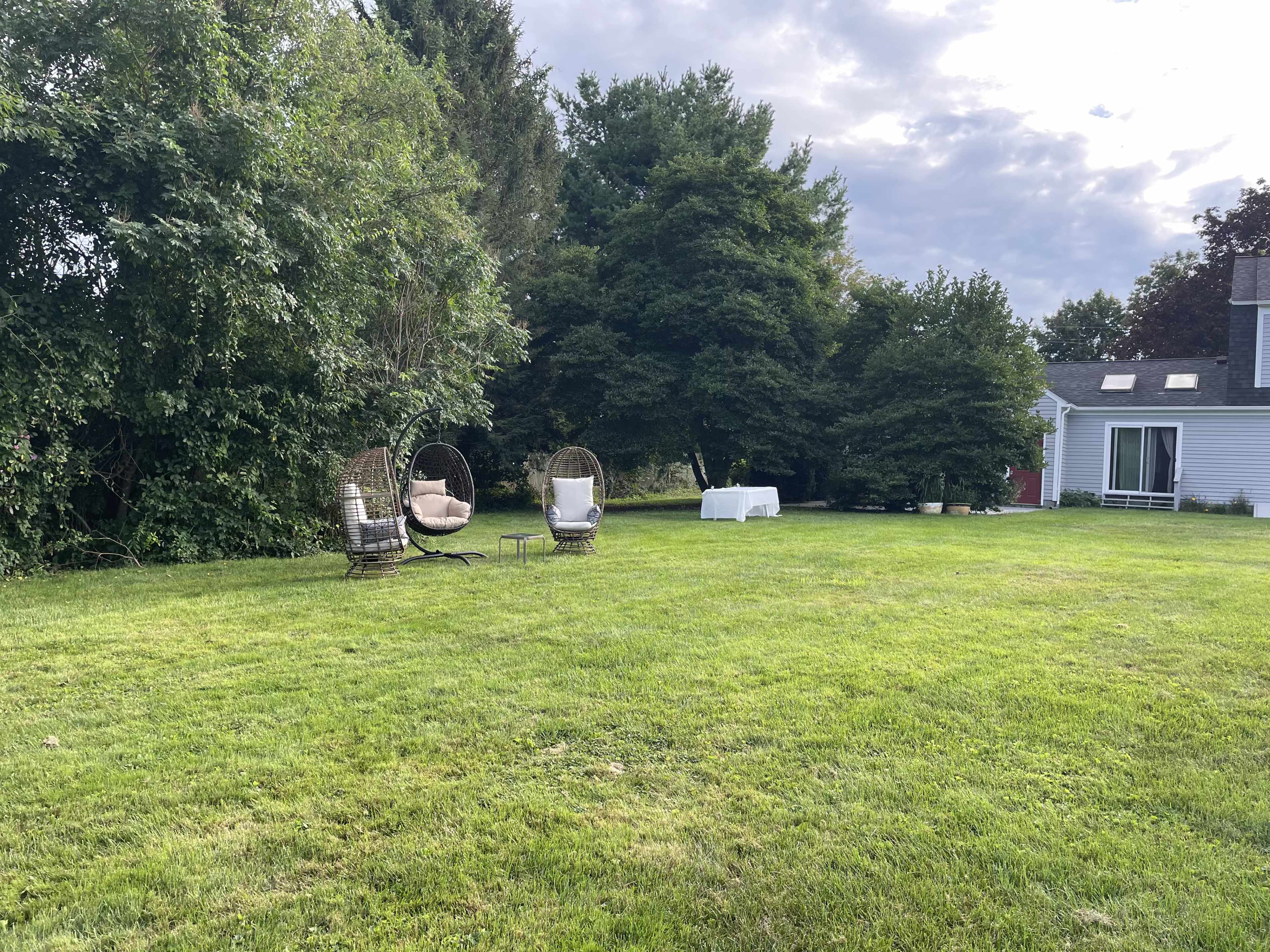 A green lawn with two hanging chairs and a small table, situated near a cluster of trees and a house in the background.