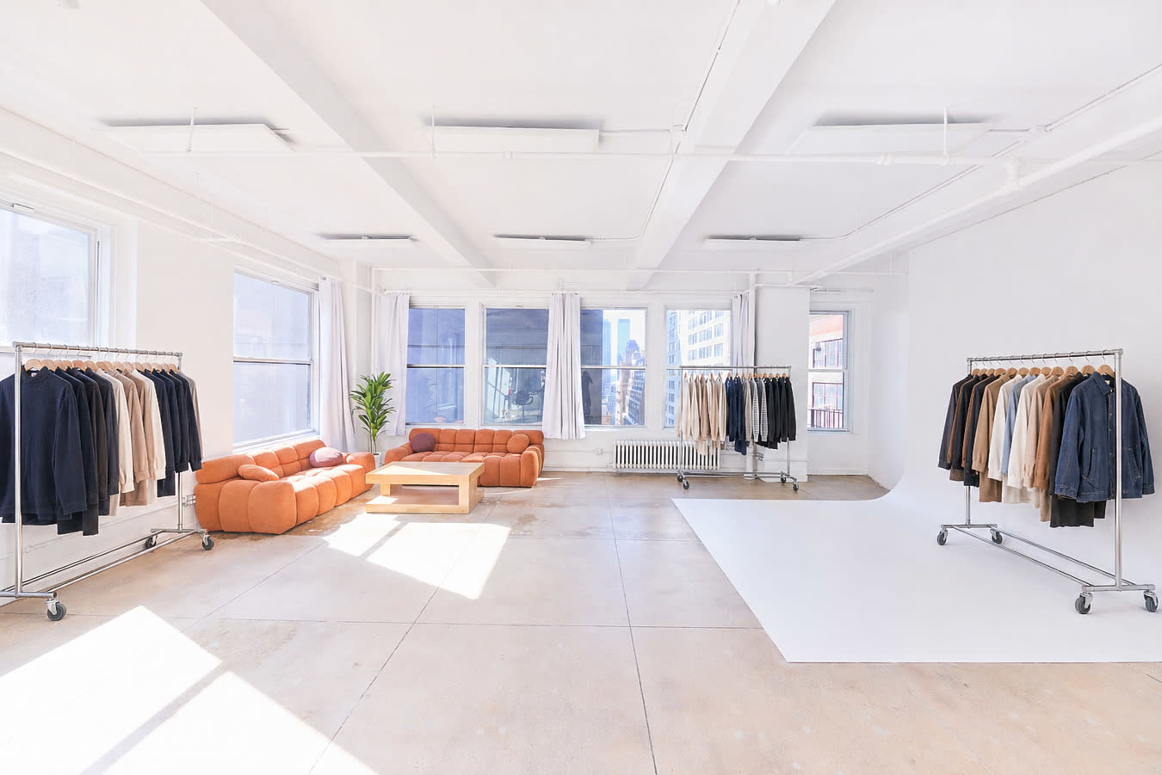 The image shows a spacious, well-lit room with large windows featuring clothing racks on either side and a sofa in the center.