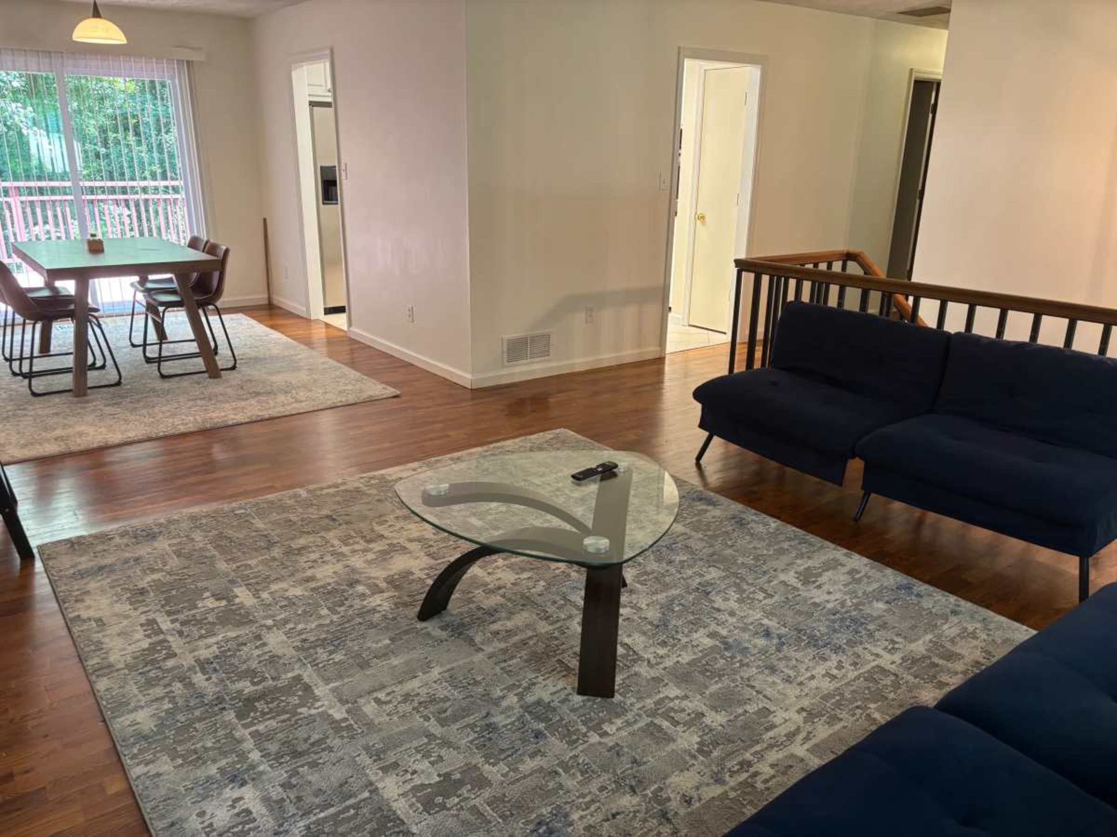 The image shows a modern living room with a blue sofa, a glass coffee table, wooden floors, and a dining area visible in the background.