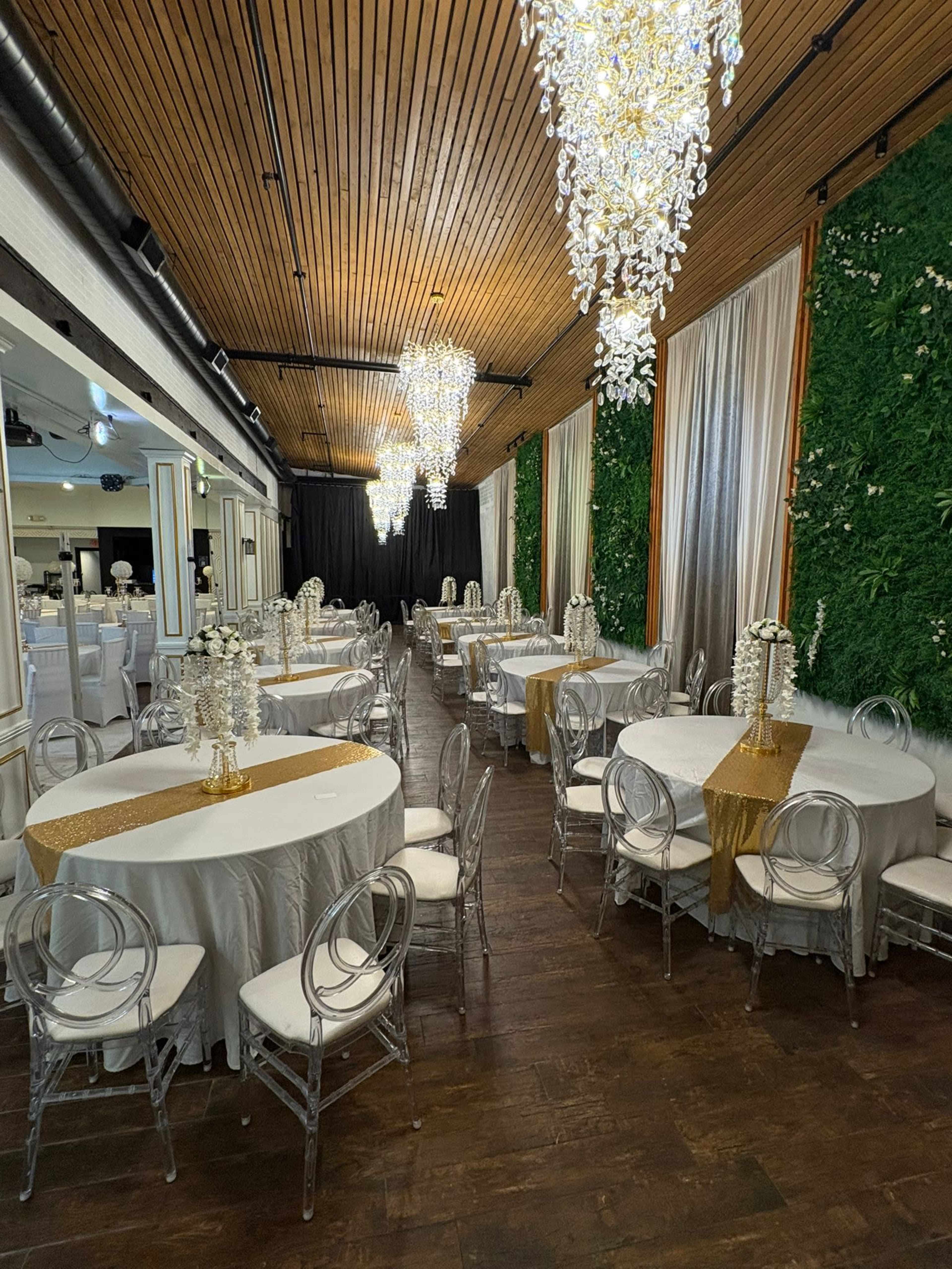 The image shows a banquet hall with circular tables covered in white tablecloths, clear ghost chairs, and elegant chandeliers hanging from a wooden ceiling, flanked by greenery-covered walls.