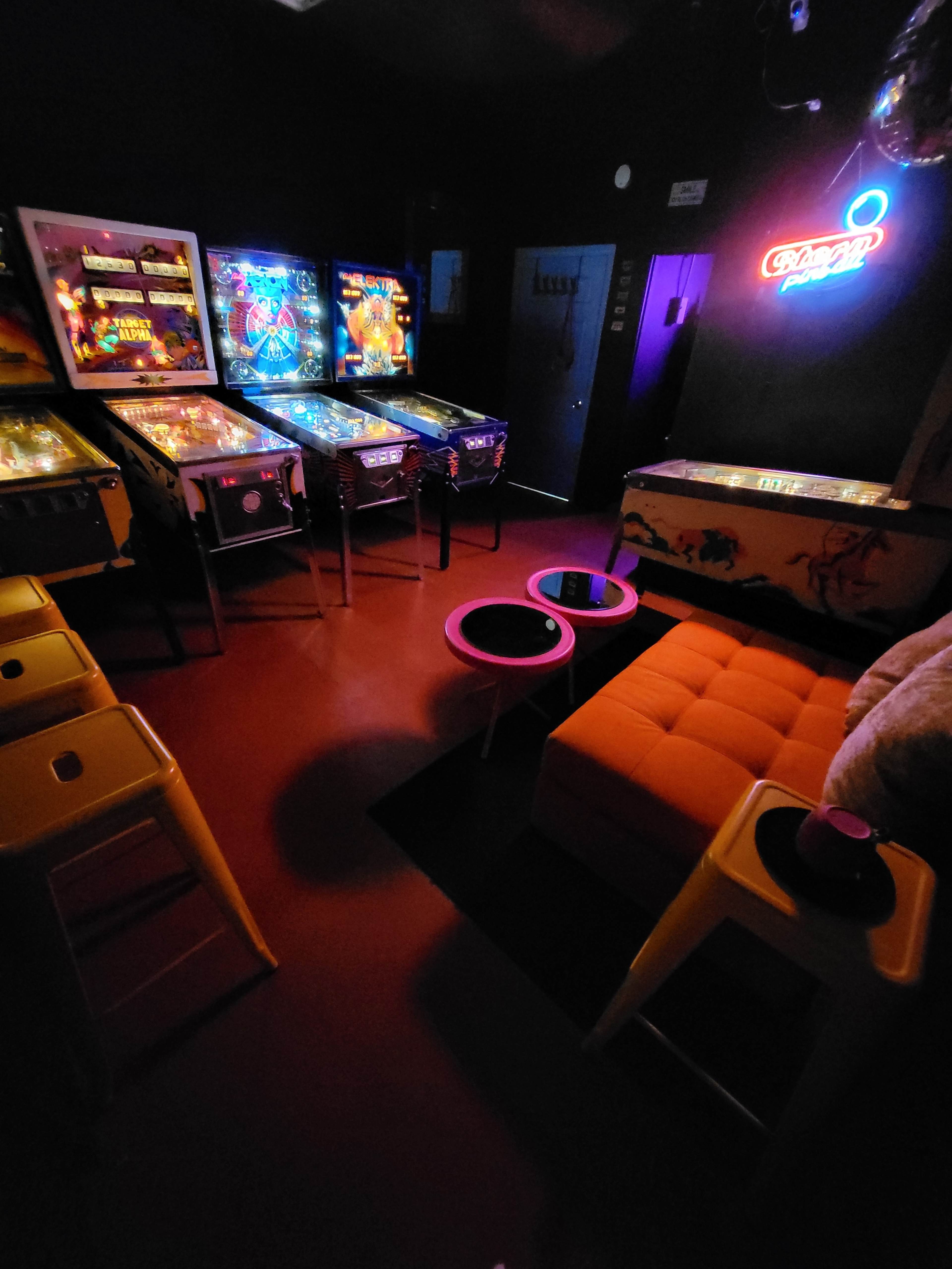 A dimly lit game room featuring several pinball machines, a bright orange couch, and colorful tables.