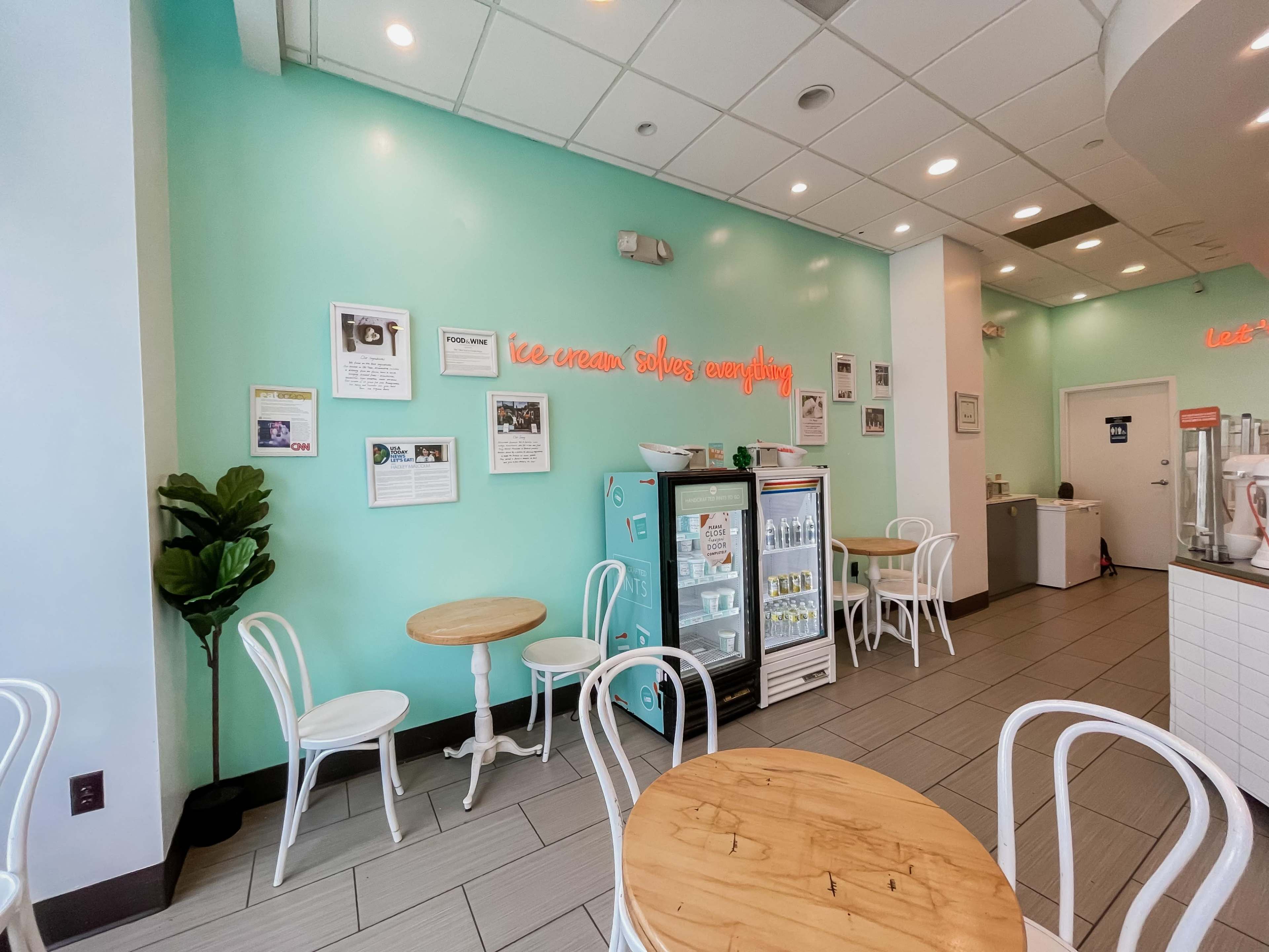 The scene shows a brightly colored ice cream shop with light teal walls, wooden tables, and white chairs, featuring a display case and various framed photos on the walls.