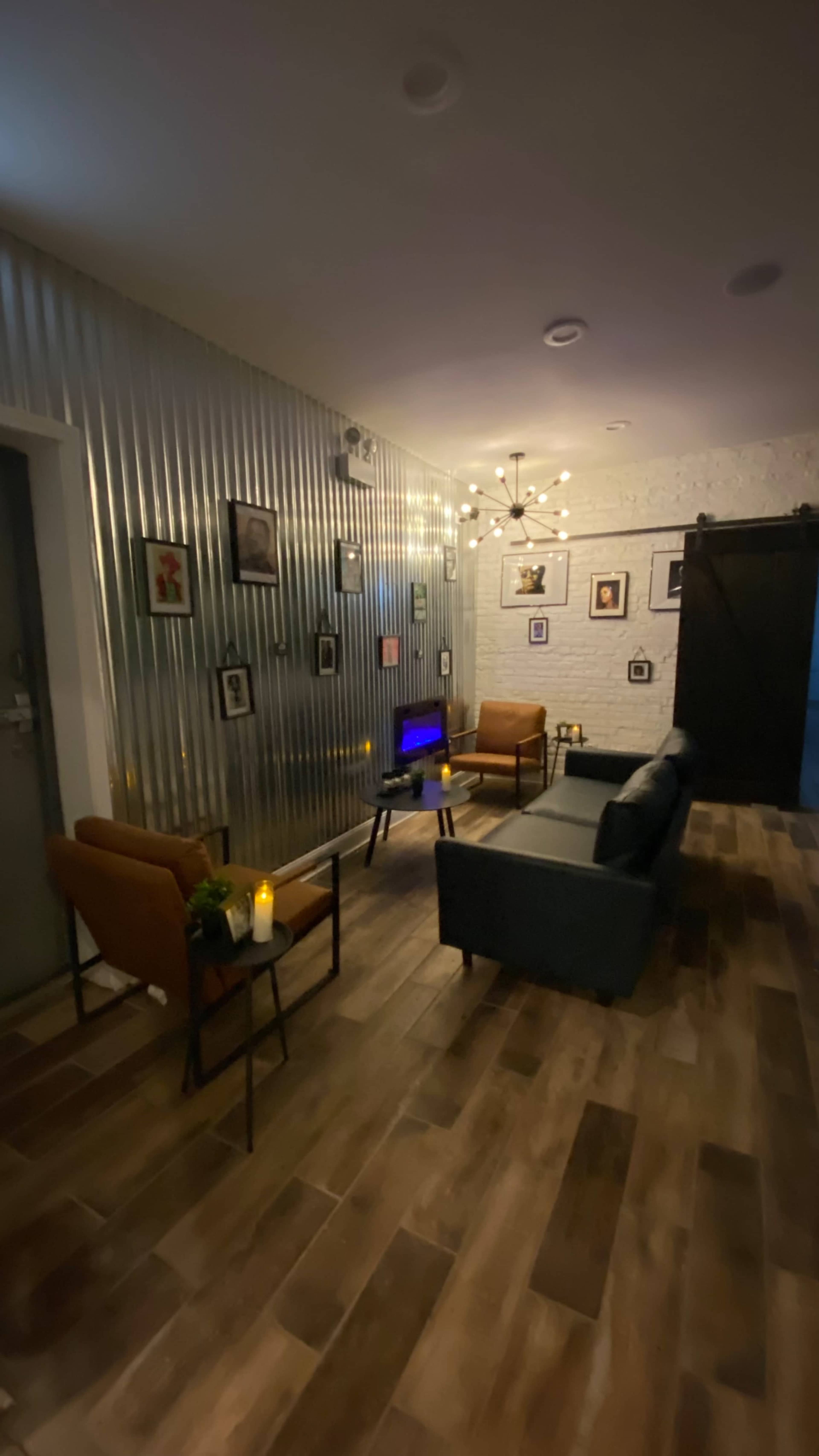 A modern living room with metal-paneled walls, wooden flooring, and a mix of chairs and a couch arranged around a television, decorated with framed artwork.