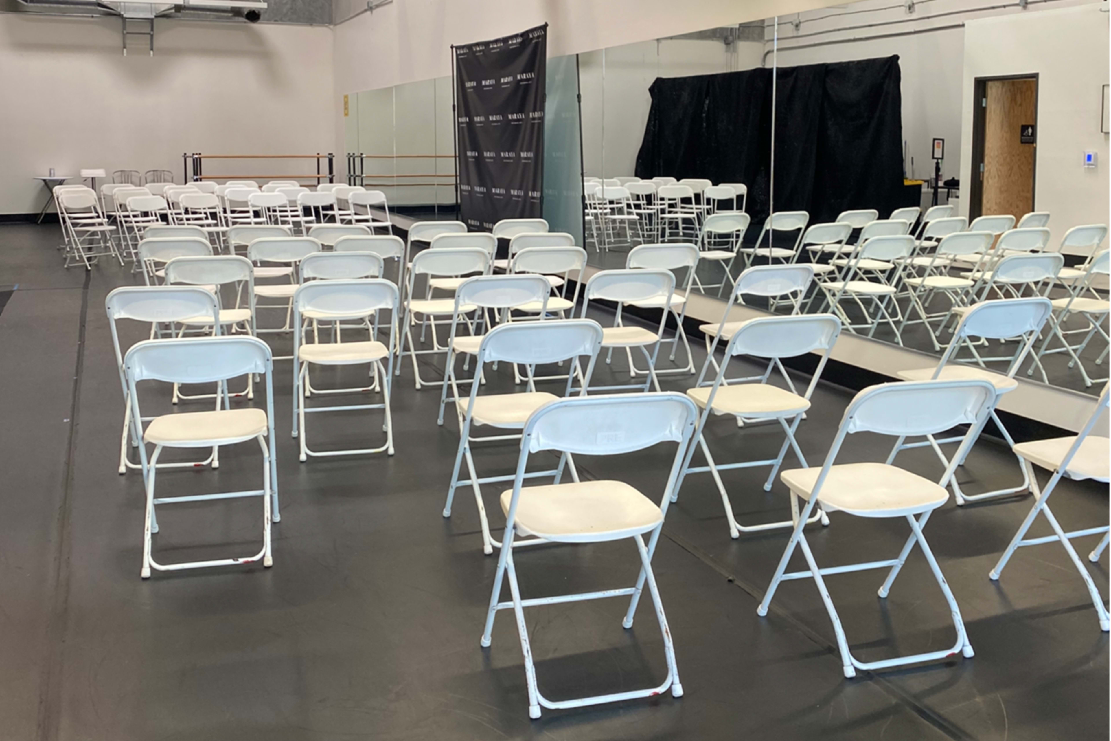 A room set up for an event, featuring rows of white folding chairs arranged in front of a mirrored wall.