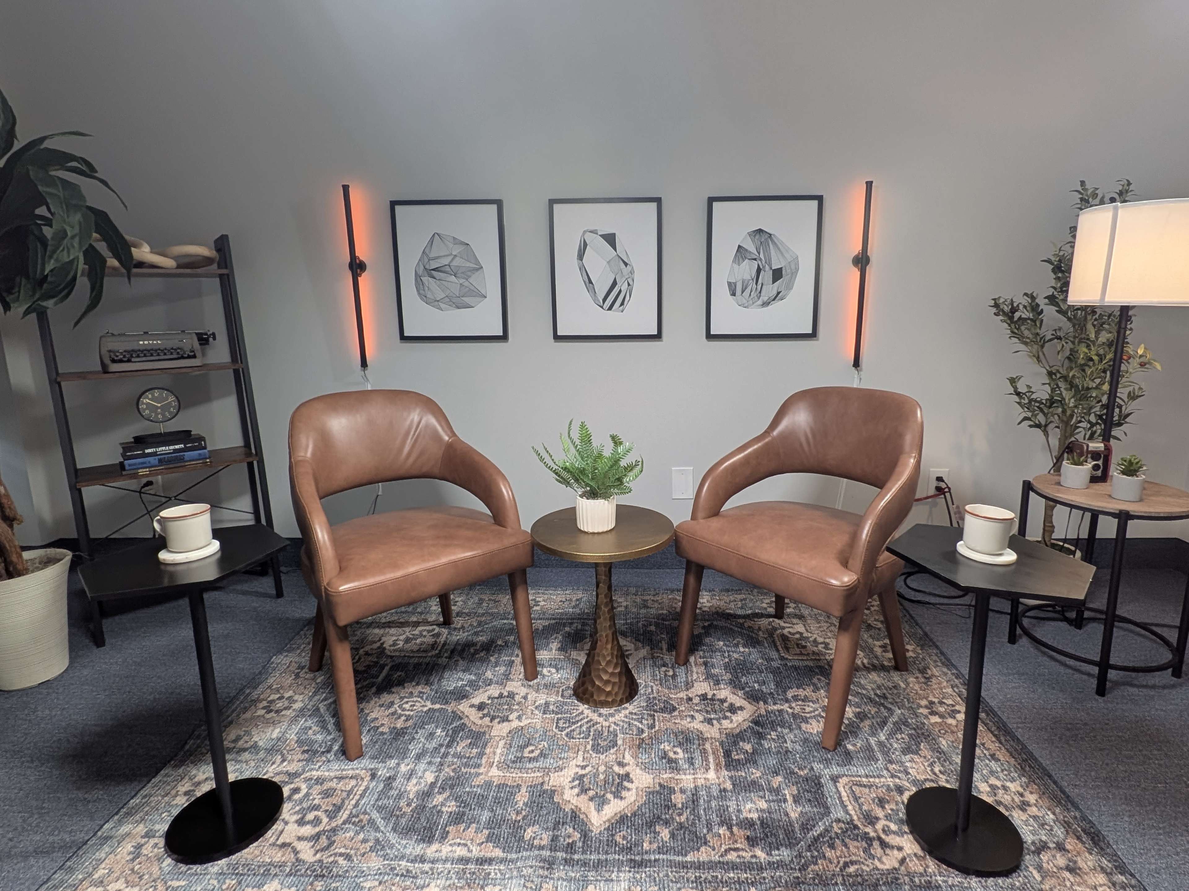 The image shows a cozy interior space featuring two brown chairs positioned around a small round table, with wall art and decorative elements in the background.