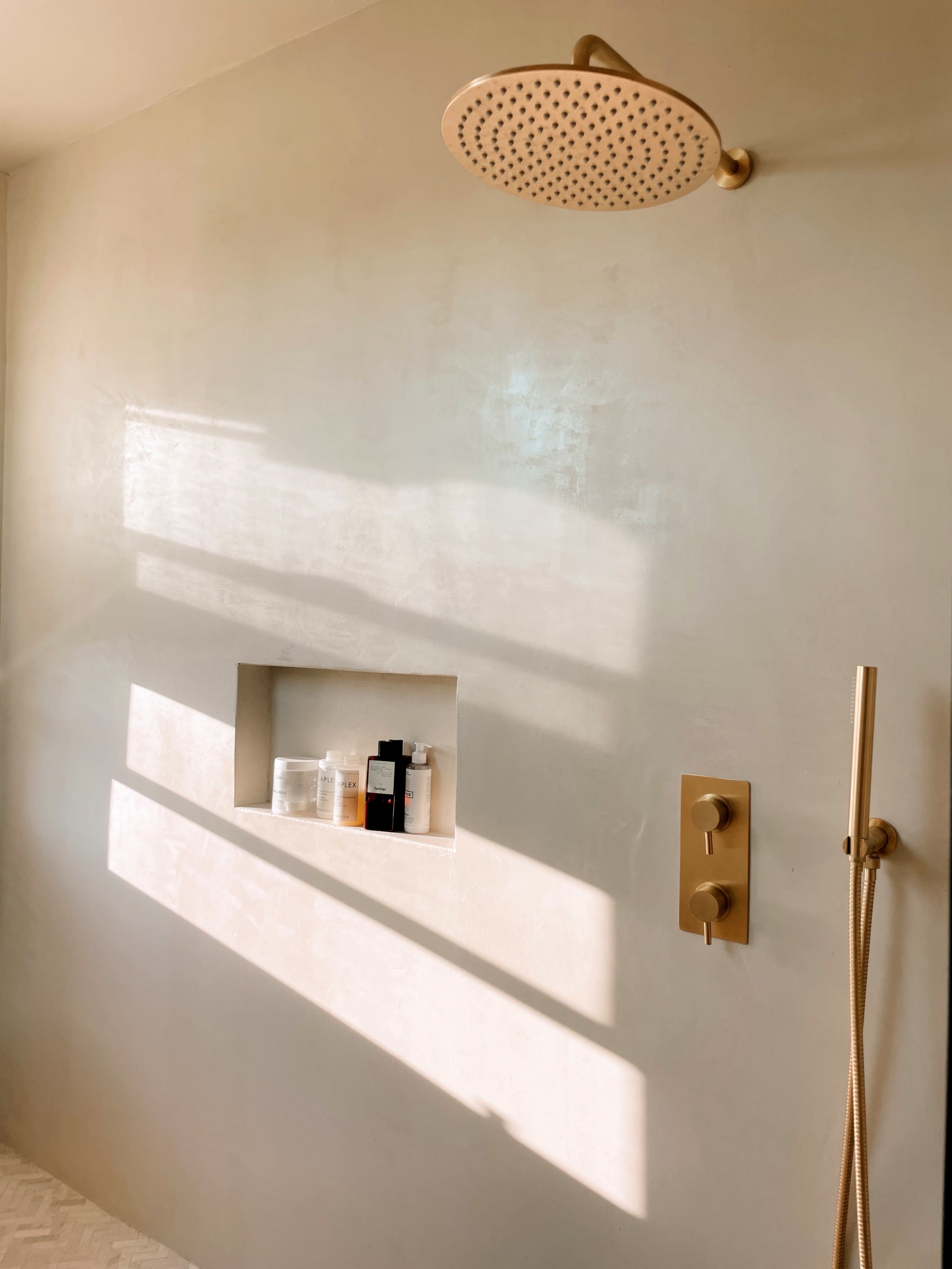 A minimalist bathroom wall with a large gold showerhead, a recessed niche containing various bottles, and light reflections casting shadows on the surface.