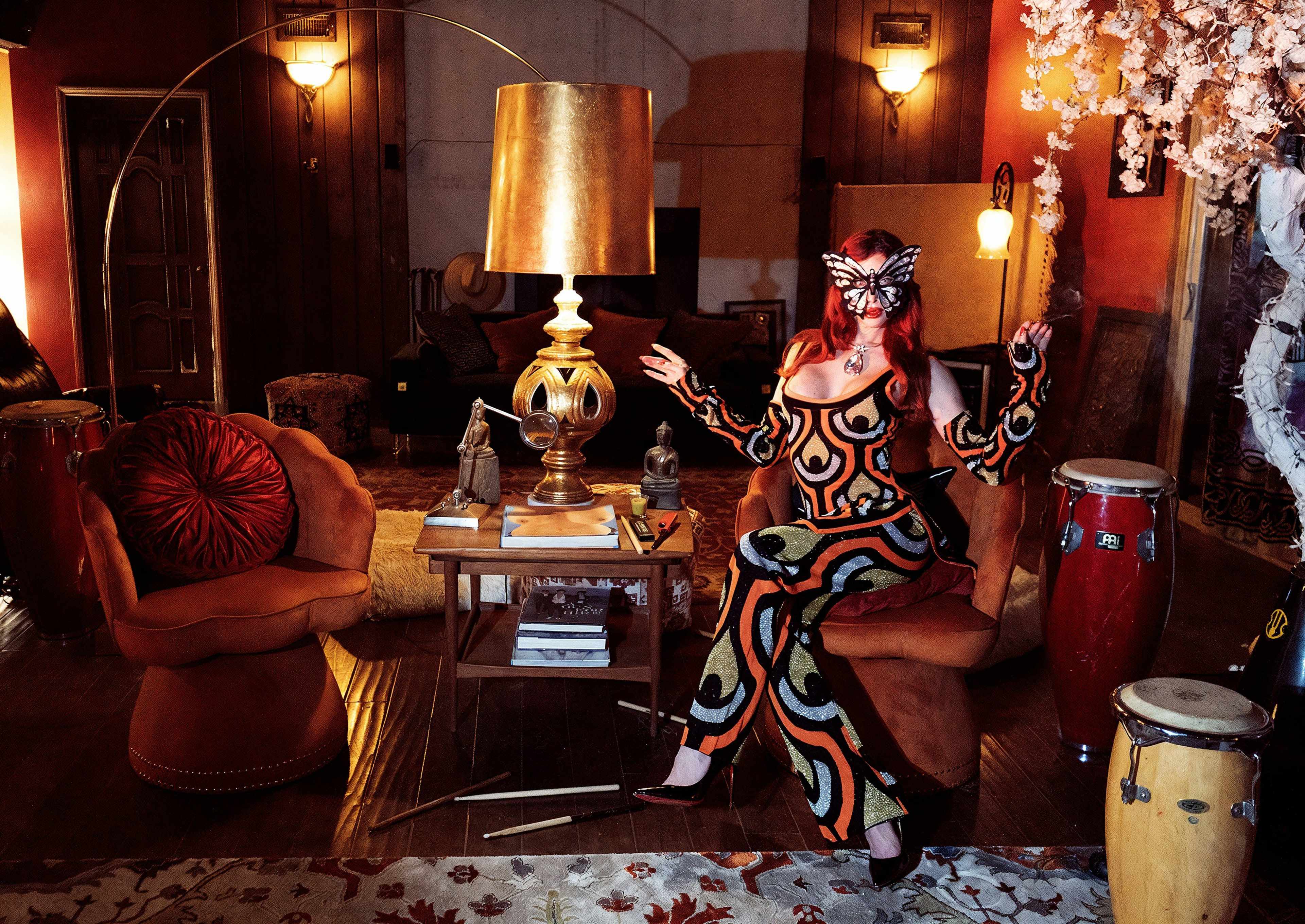 A woman in a colorful patterned outfit poses in a room decorated with a large gold lamp, musical instruments, and various furniture.