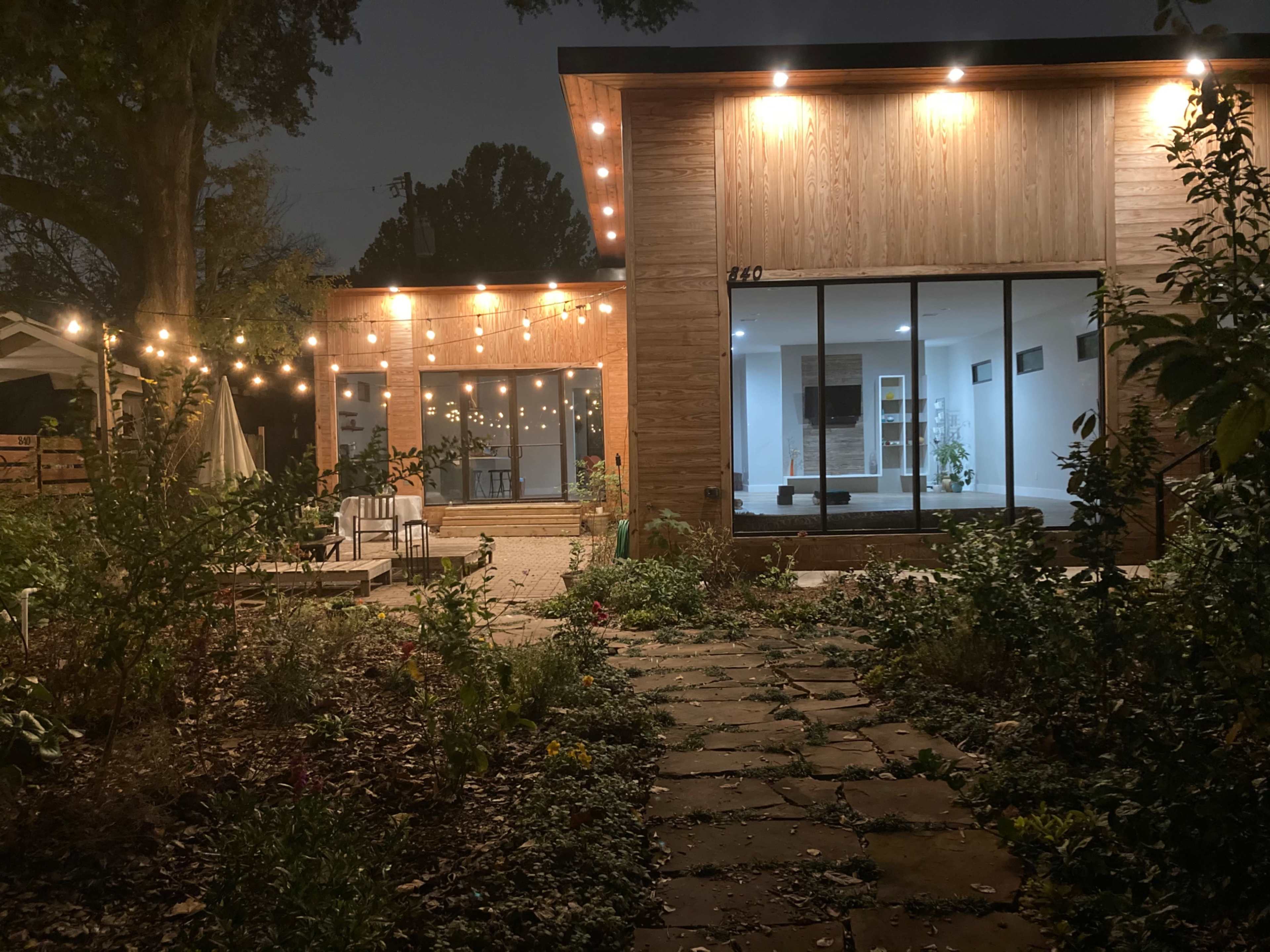 A modern house at night, illuminated by string lights, with a landscaped garden path leading to it.