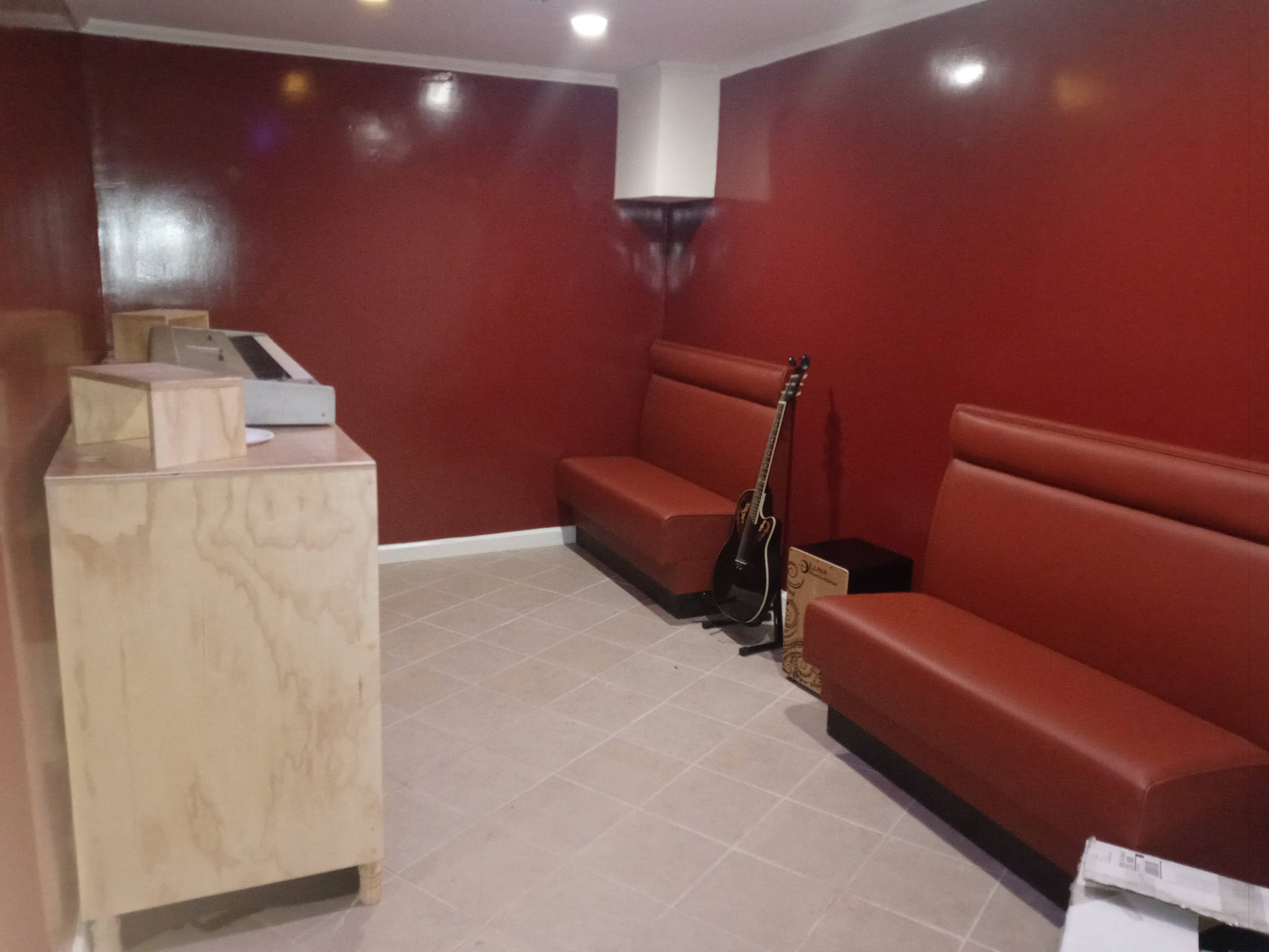 The image shows a small, sparsely furnished room with red walls, featuring two brown leather benches and a guitar leaning against the wall.