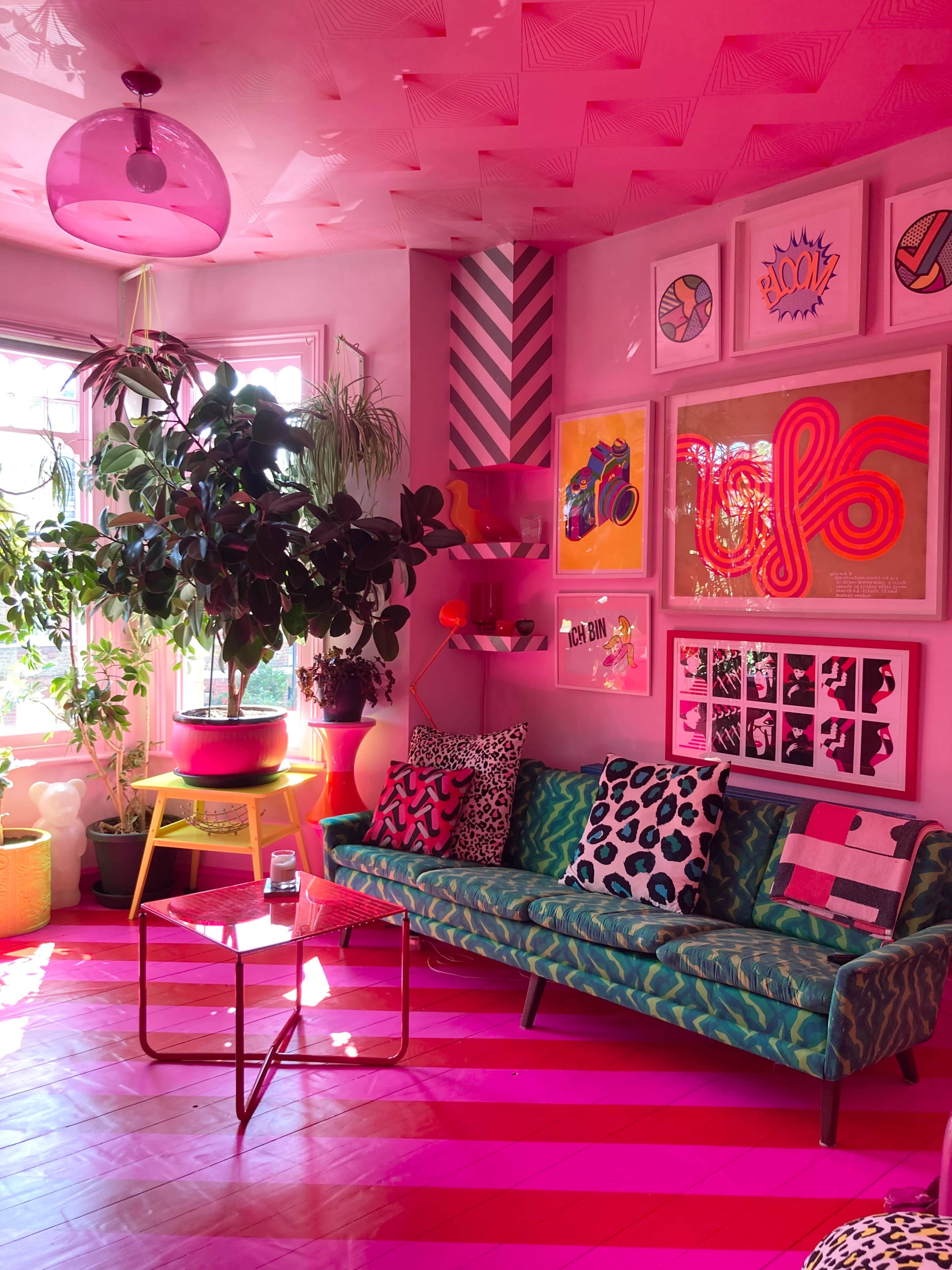 The image shows a brightly decorated living room with a pink color scheme, featuring a green sofa, various plants, and colorful artwork on the walls.