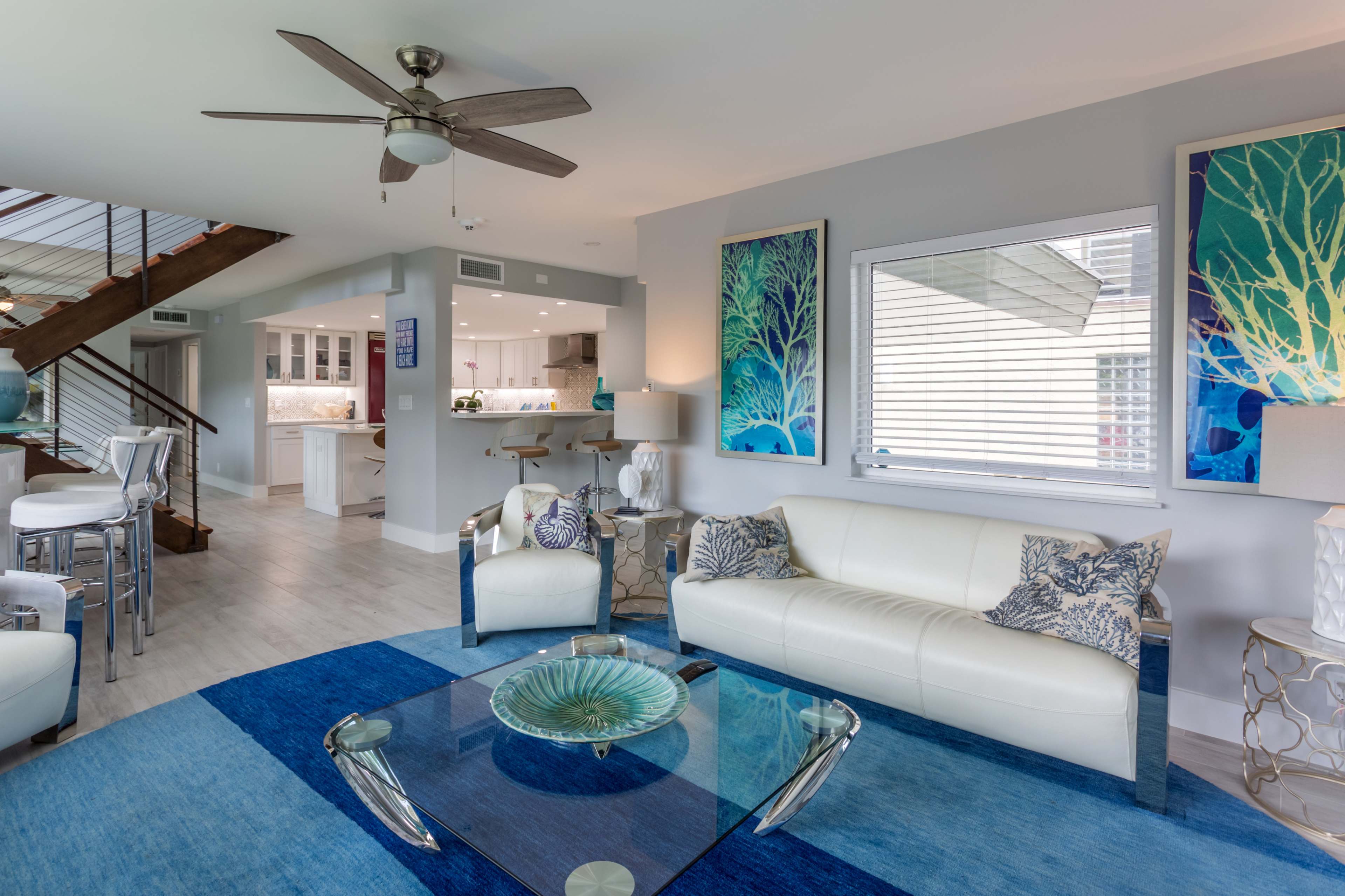 A modern living room with a white sofa, a glass coffee table, and blue artwork on the walls, overlooking a kitchen area.