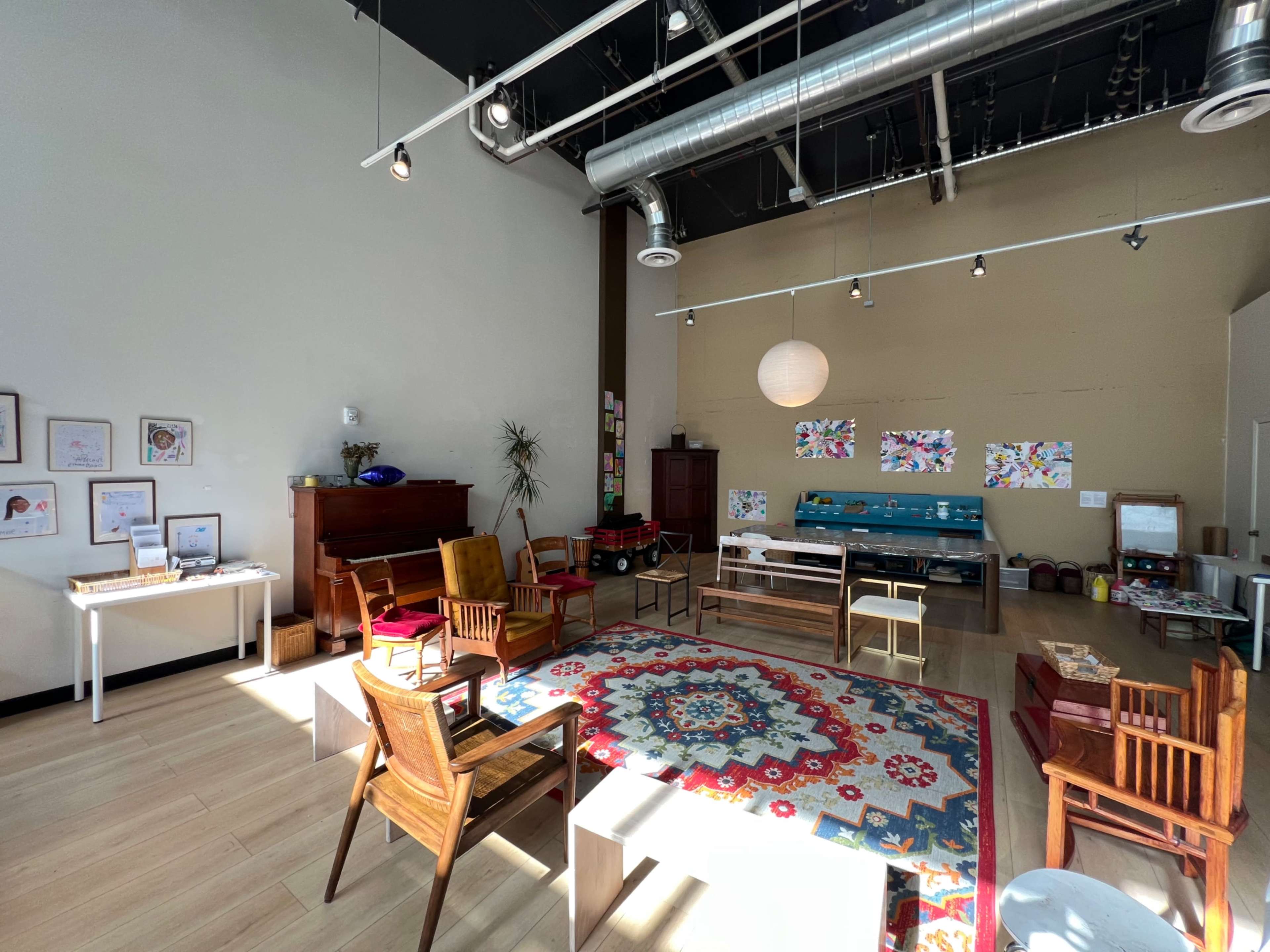 The image shows a spacious room with wooden furniture, including chairs and tables, arranged around a colorful rug, with artwork displayed on the walls.