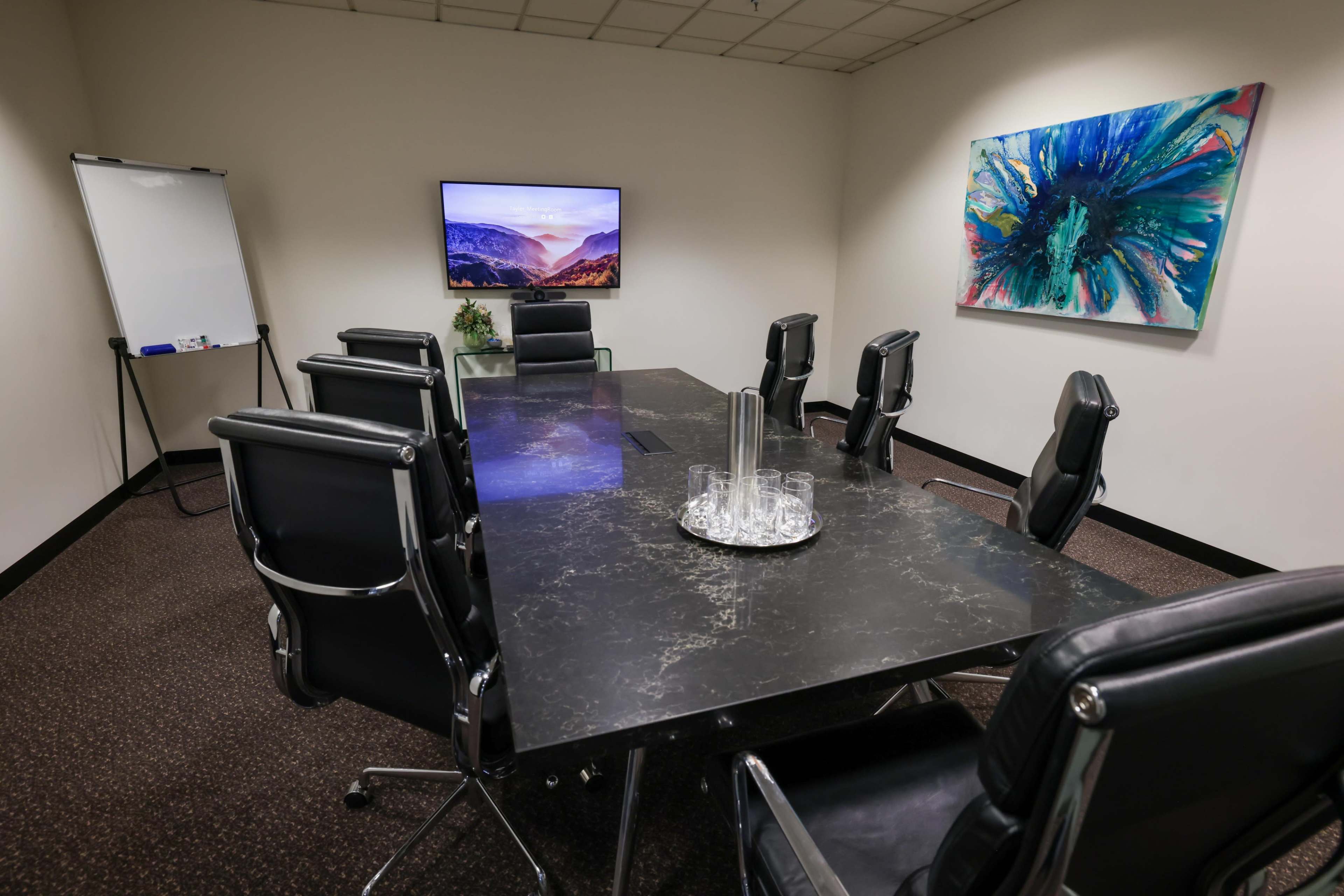 The image shows a modern conference room with a large table surrounded by black leather chairs, a whiteboard on one side, and an abstract painting on the wall.