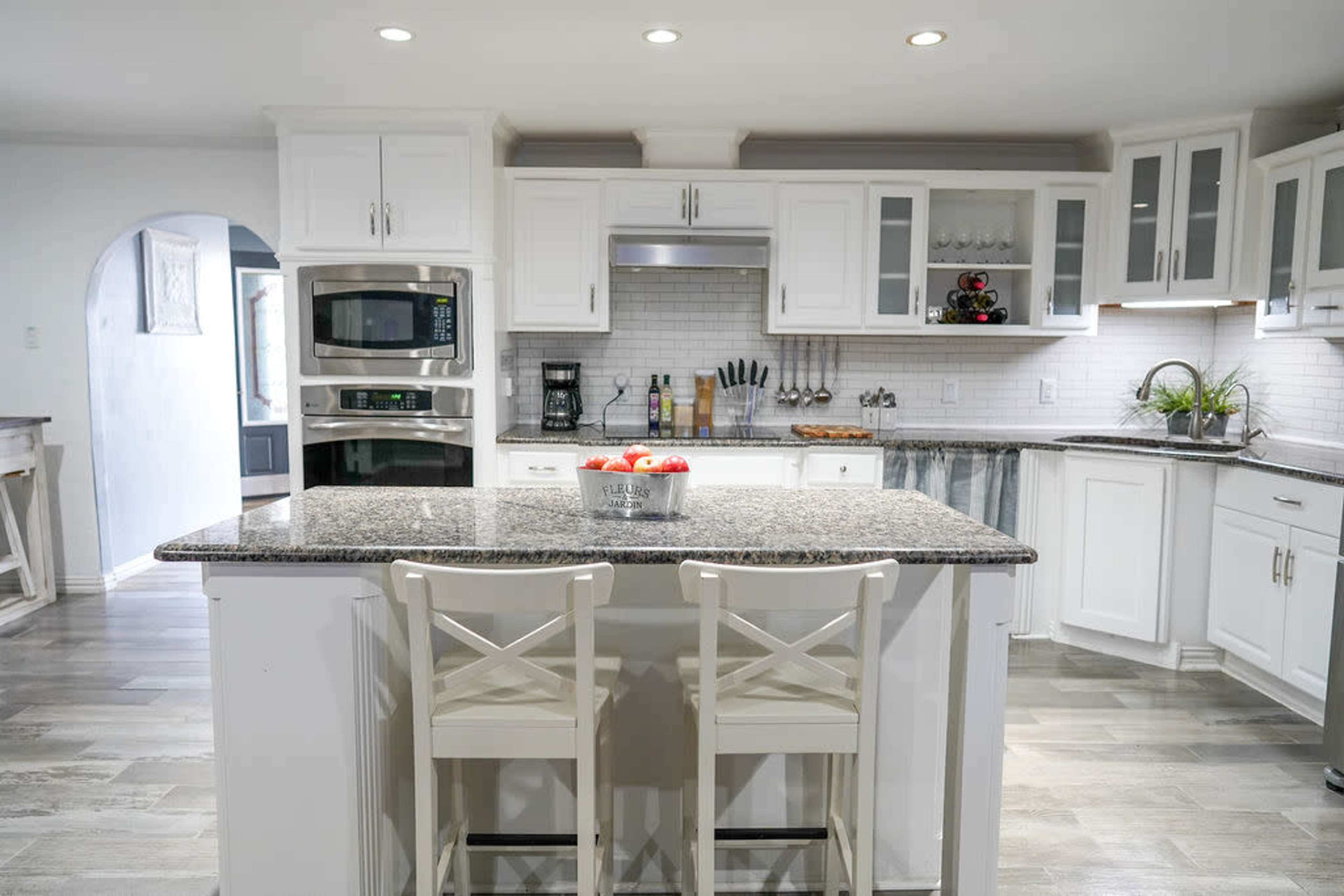 A modern kitchen features a central island with two chairs, white cabinets, stainless steel appliances, and granite countertops.