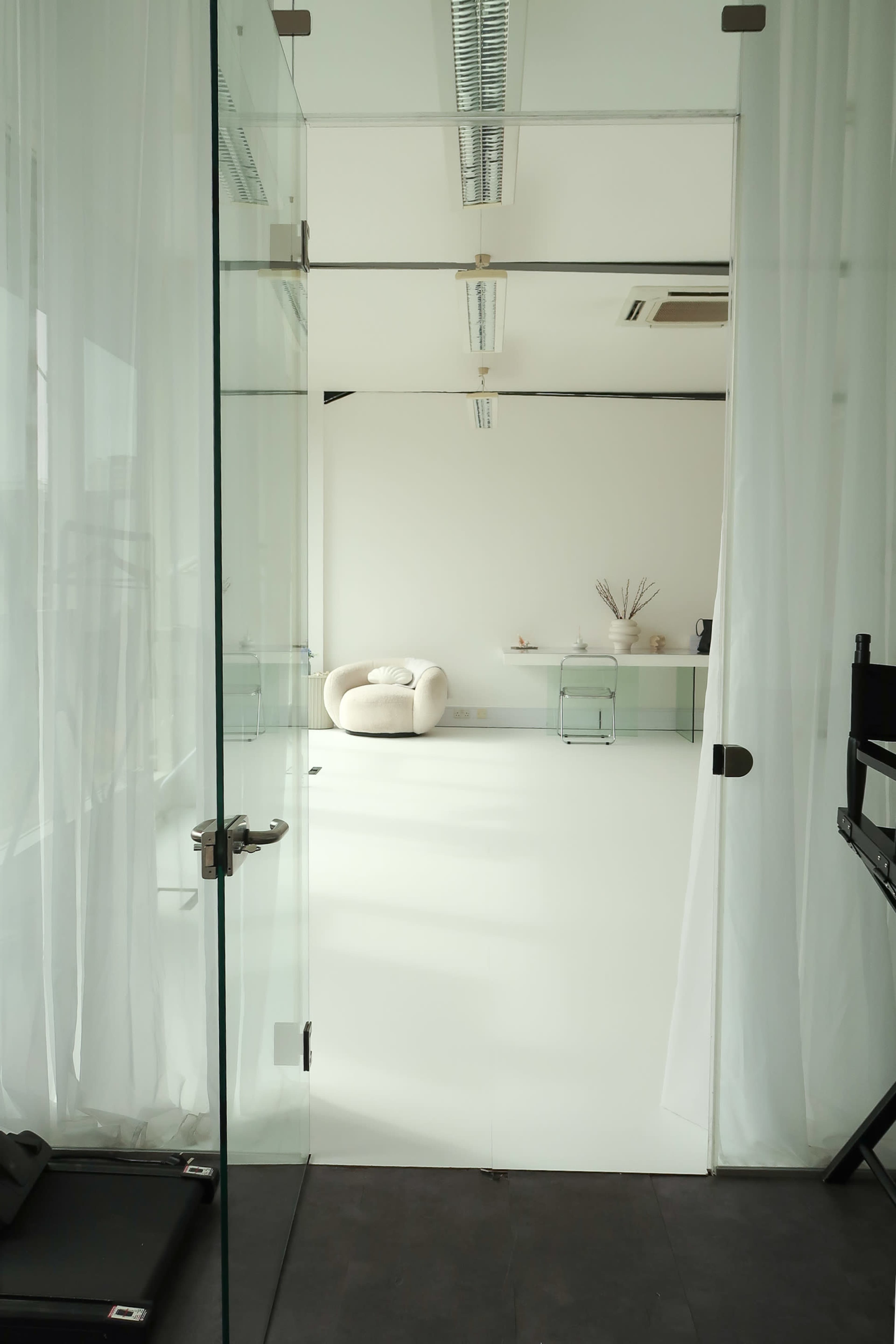 A glass door leads into a bright, minimalistic room featuring a white floor, a large armchair, and a small table against a plain wall.