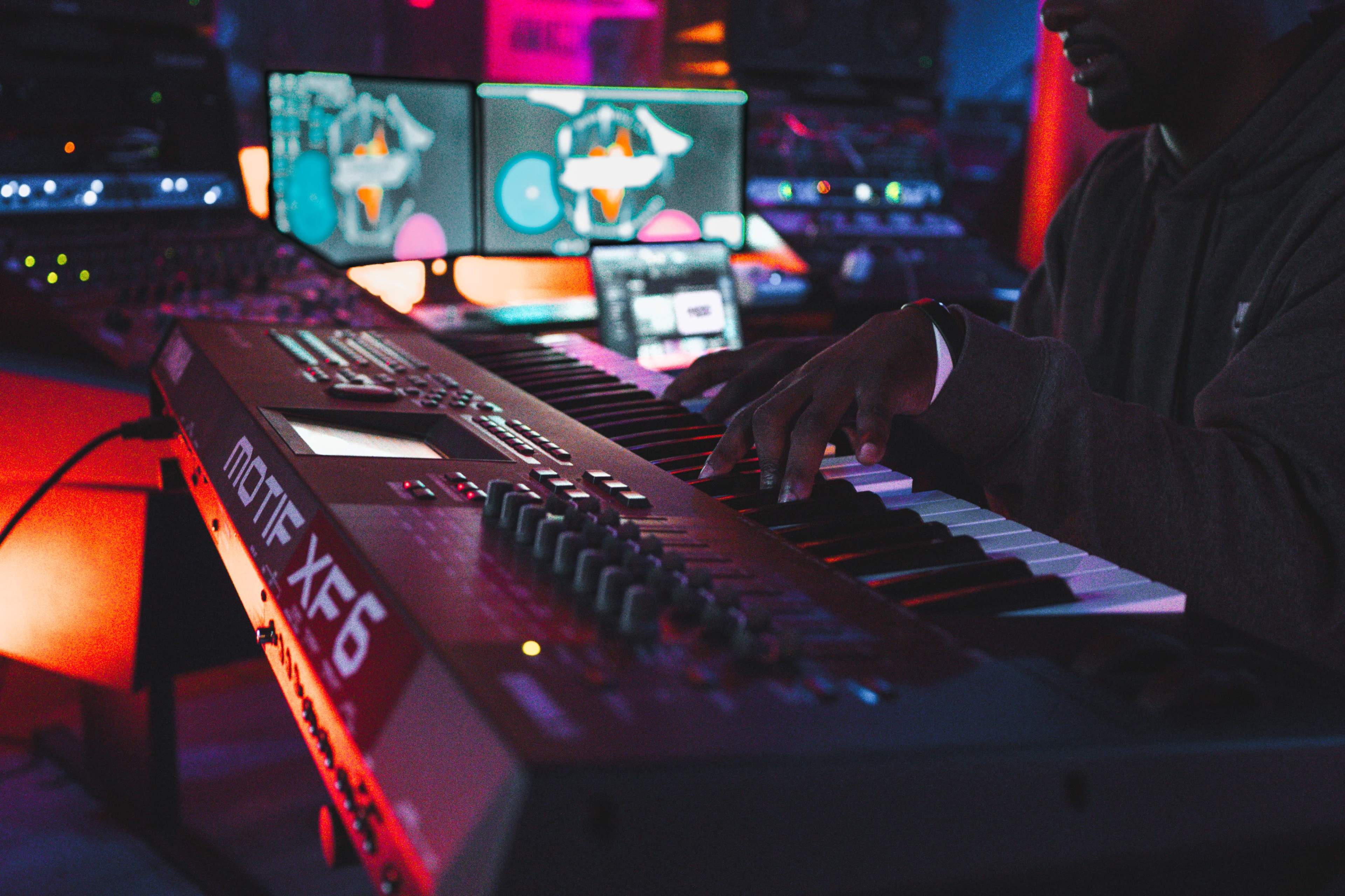A musician plays a keyboard in a dimly lit studio filled with electronic equipment and screens displaying audio software.