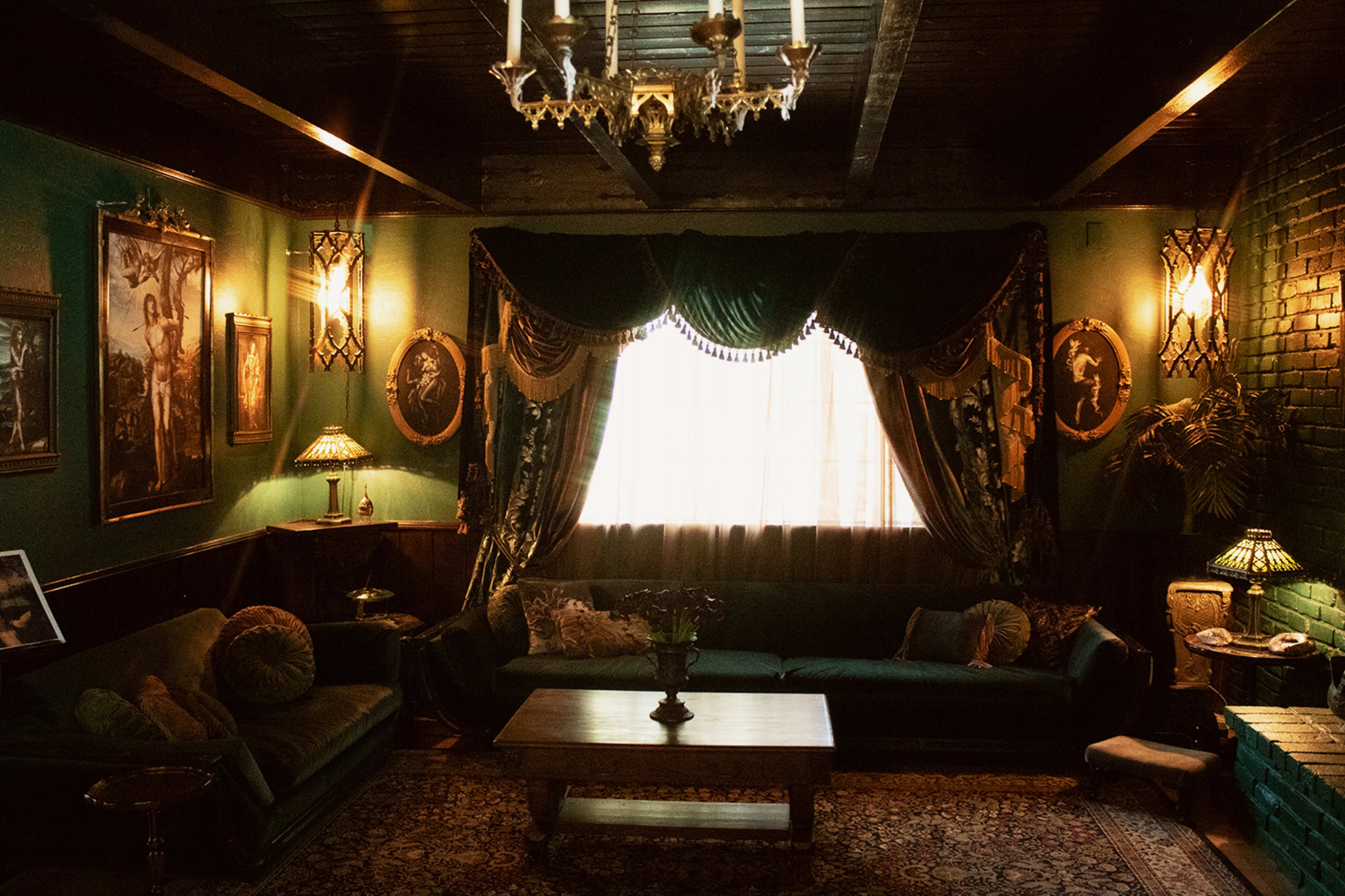 The image shows a dimly lit living room with green walls, a large window draped with ornate curtains, and vintage furniture including a dark sofa and a wooden coffee table.