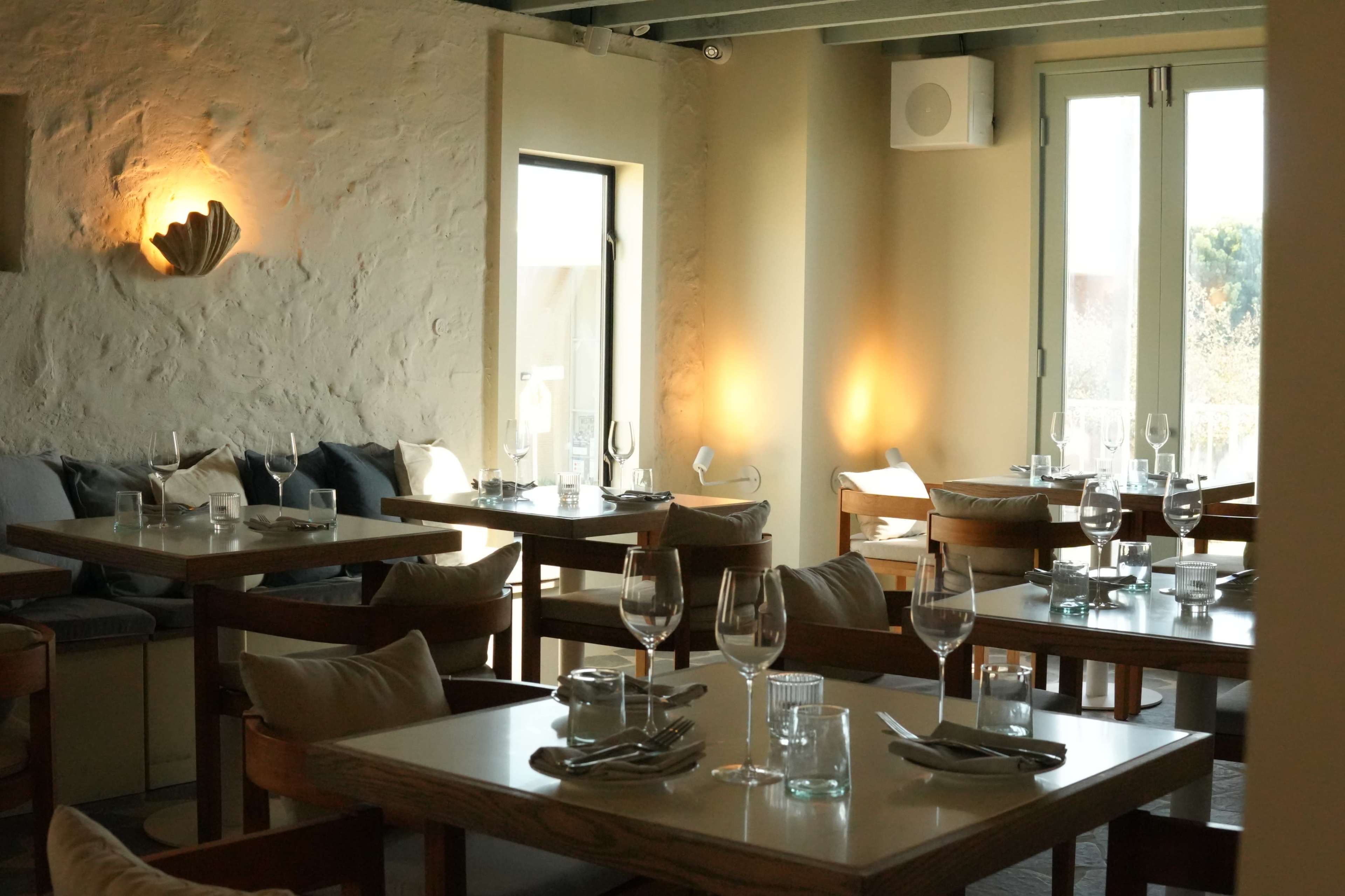 The image shows an elegantly designed restaurant interior with wooden tables, glassware, and soft seating arrangements near large windows.