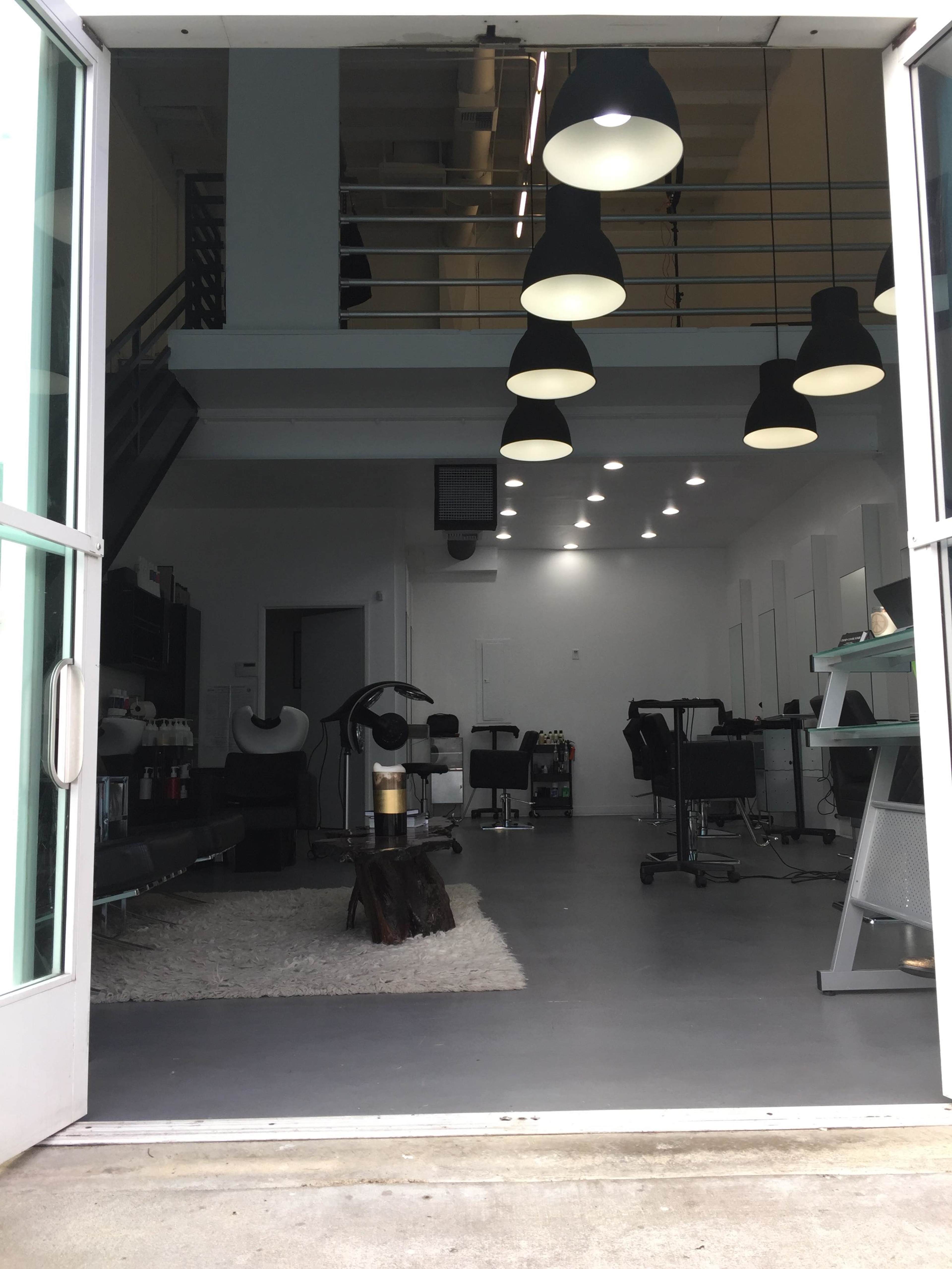 The interior of a modern salon featuring several styling stations, a waiting area, and various overhead lights.