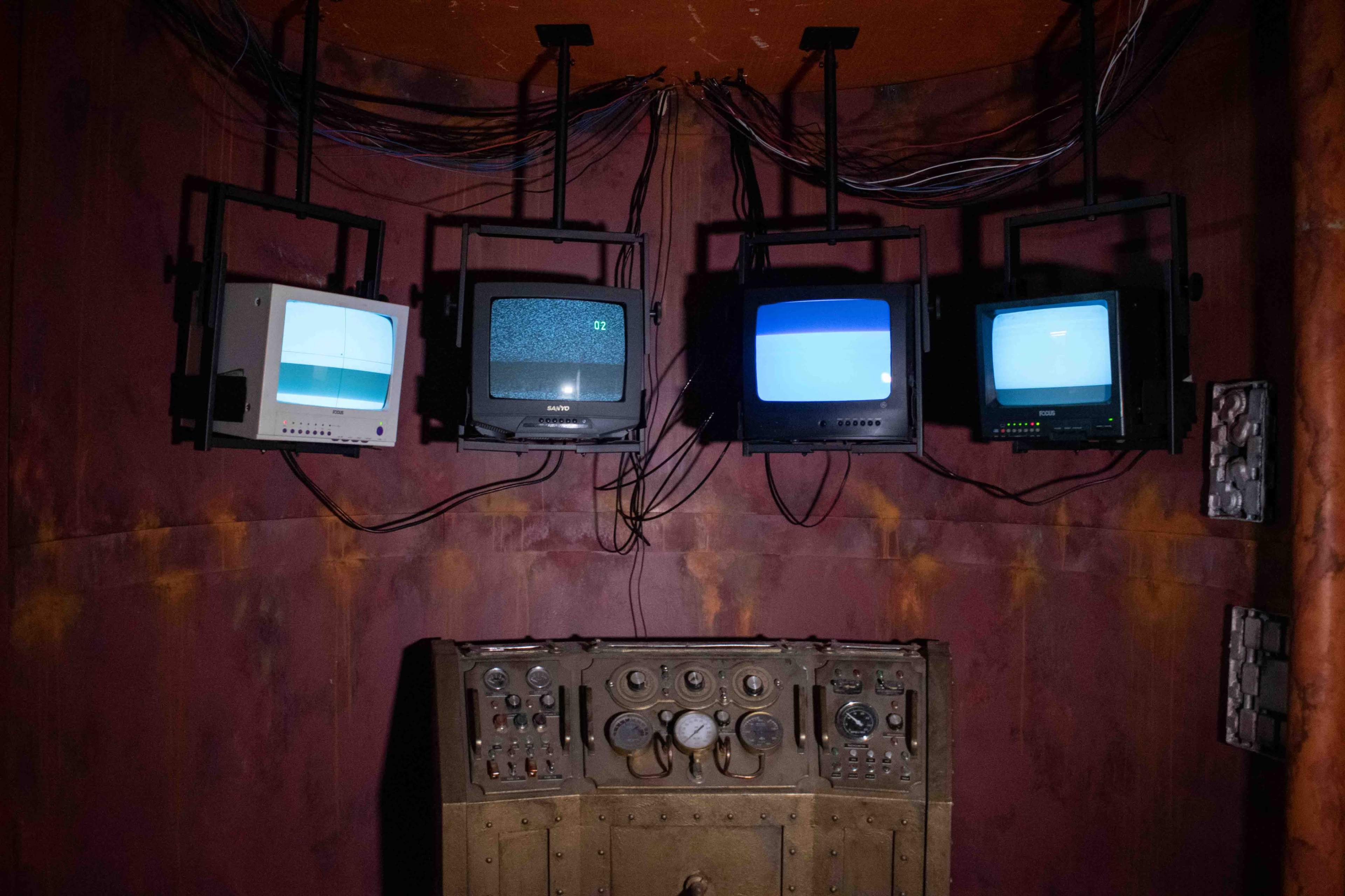 A dimly lit room with five old televisions mounted on the wall, displaying static, above a control panel with various knobs and dials.