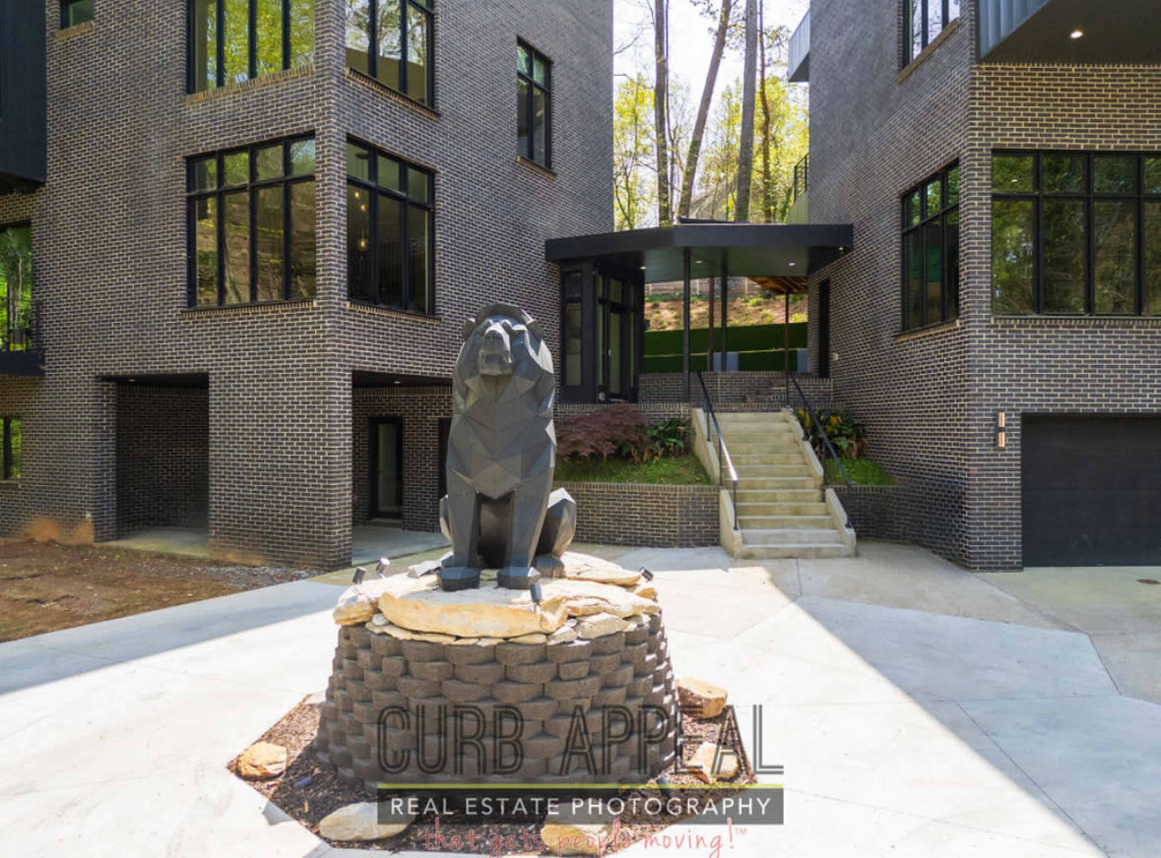 A geometric lion sculpture sits on a stone pedestal in front of a contemporary, multi-level brick building with large windows.