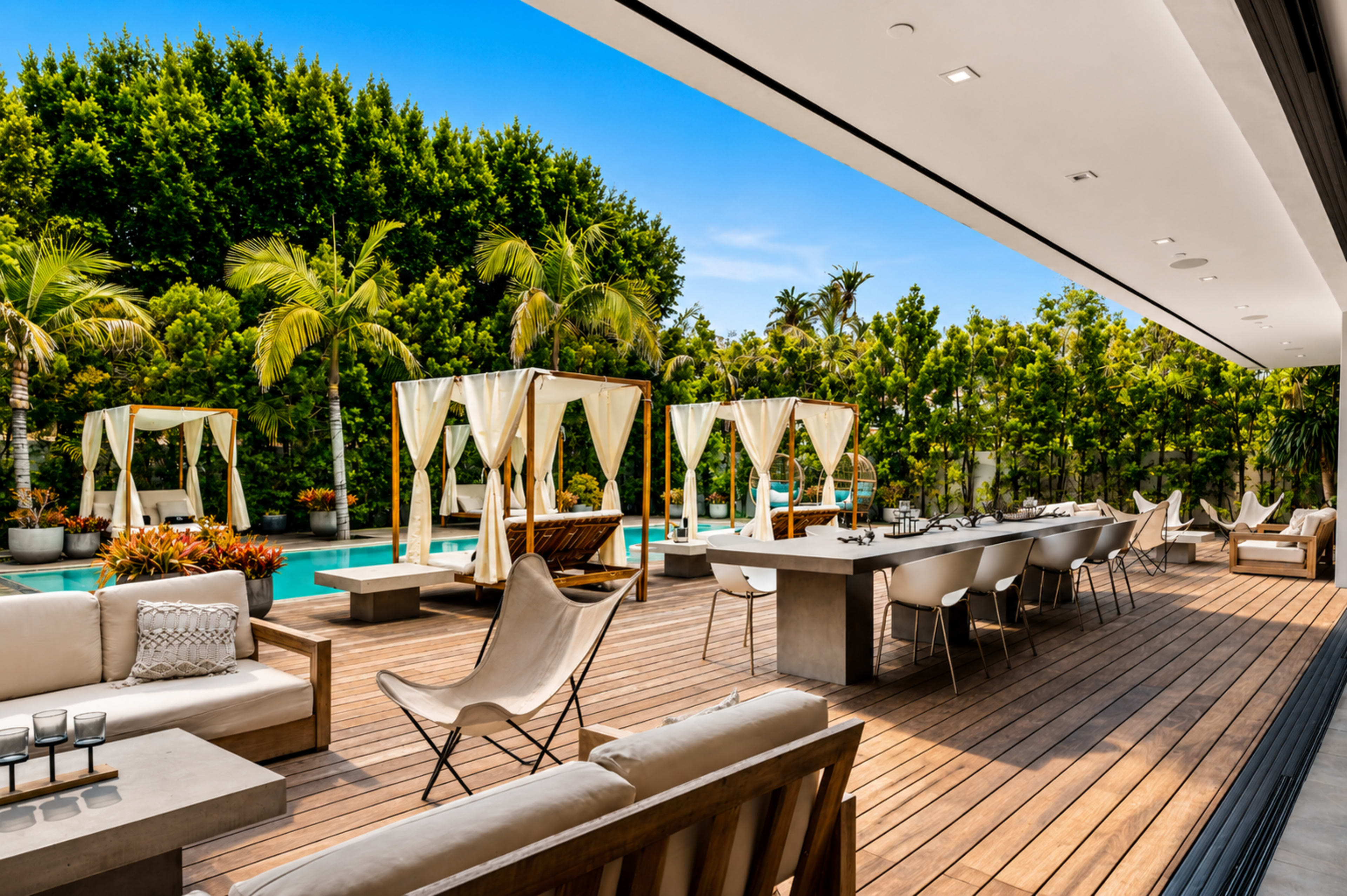 The image shows a modern outdoor patio area with seating, a dining table, and cabanas overlooking a swimming pool surrounded by lush greenery.