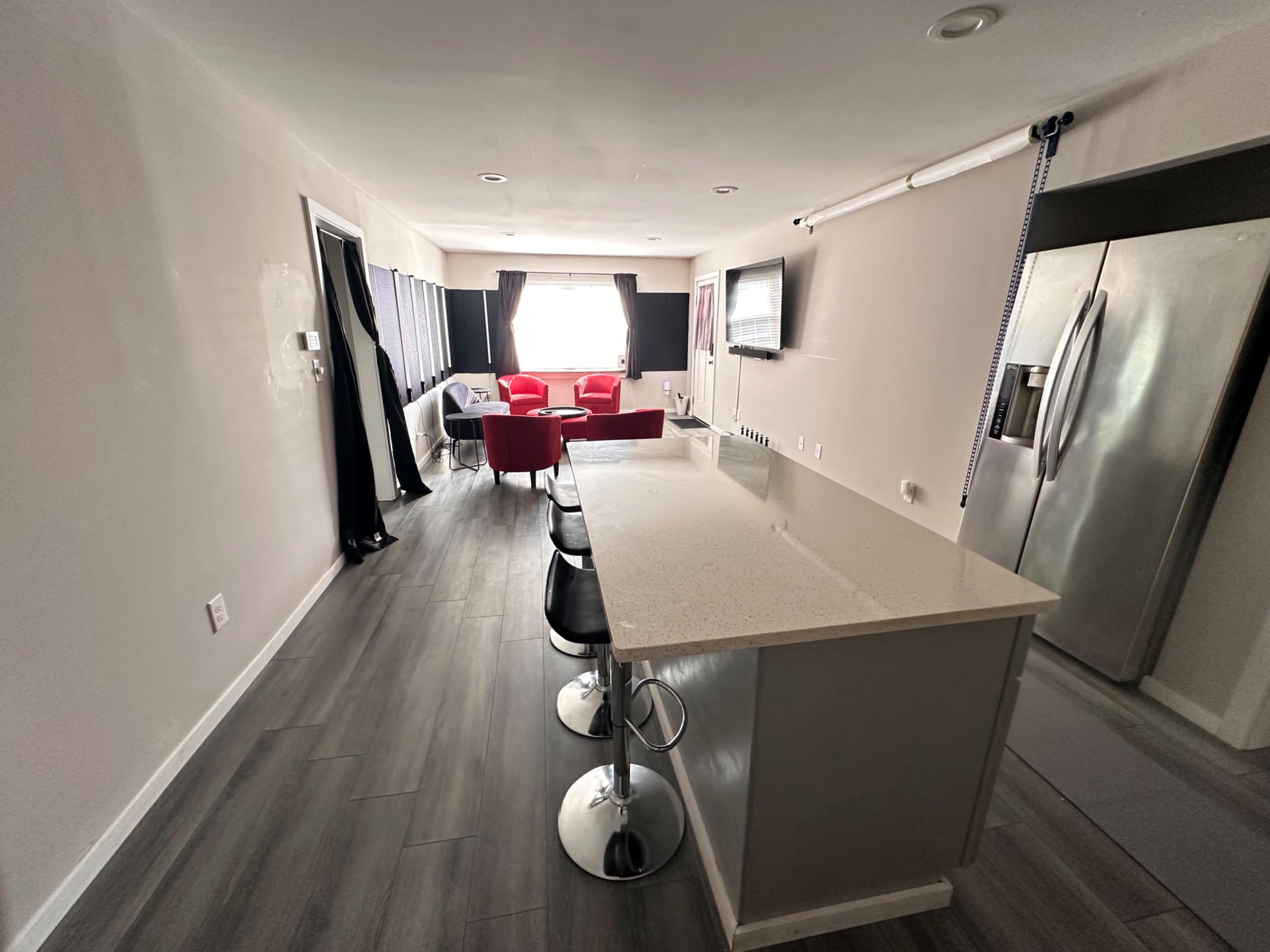 The image shows a modern kitchen area with a countertop bar and seating, leading into a living space furnished with red chairs and a television.