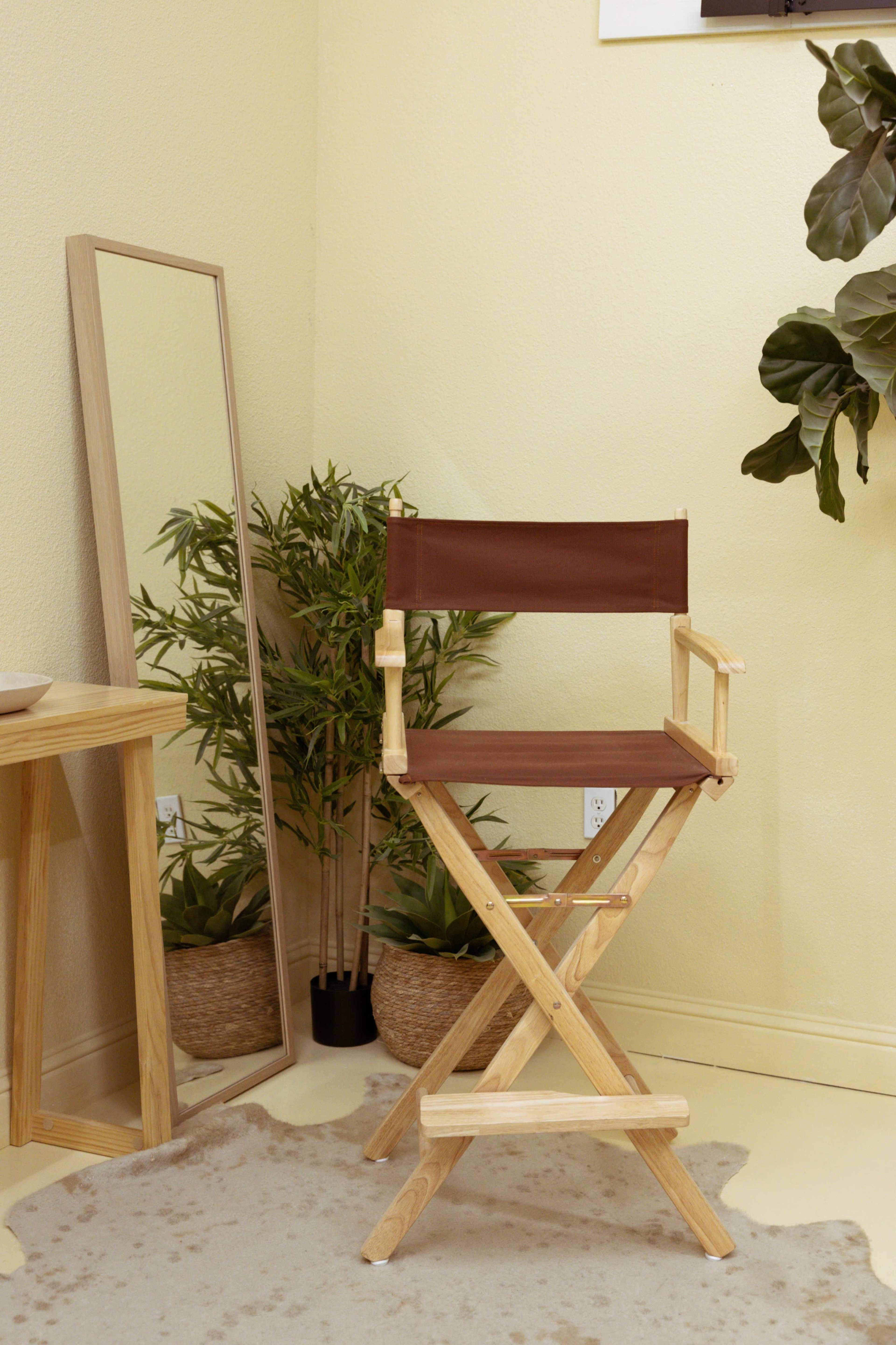 A wooden director's chair sits in a corner next to a tall mirror and a potted plant, against a pale yellow wall.