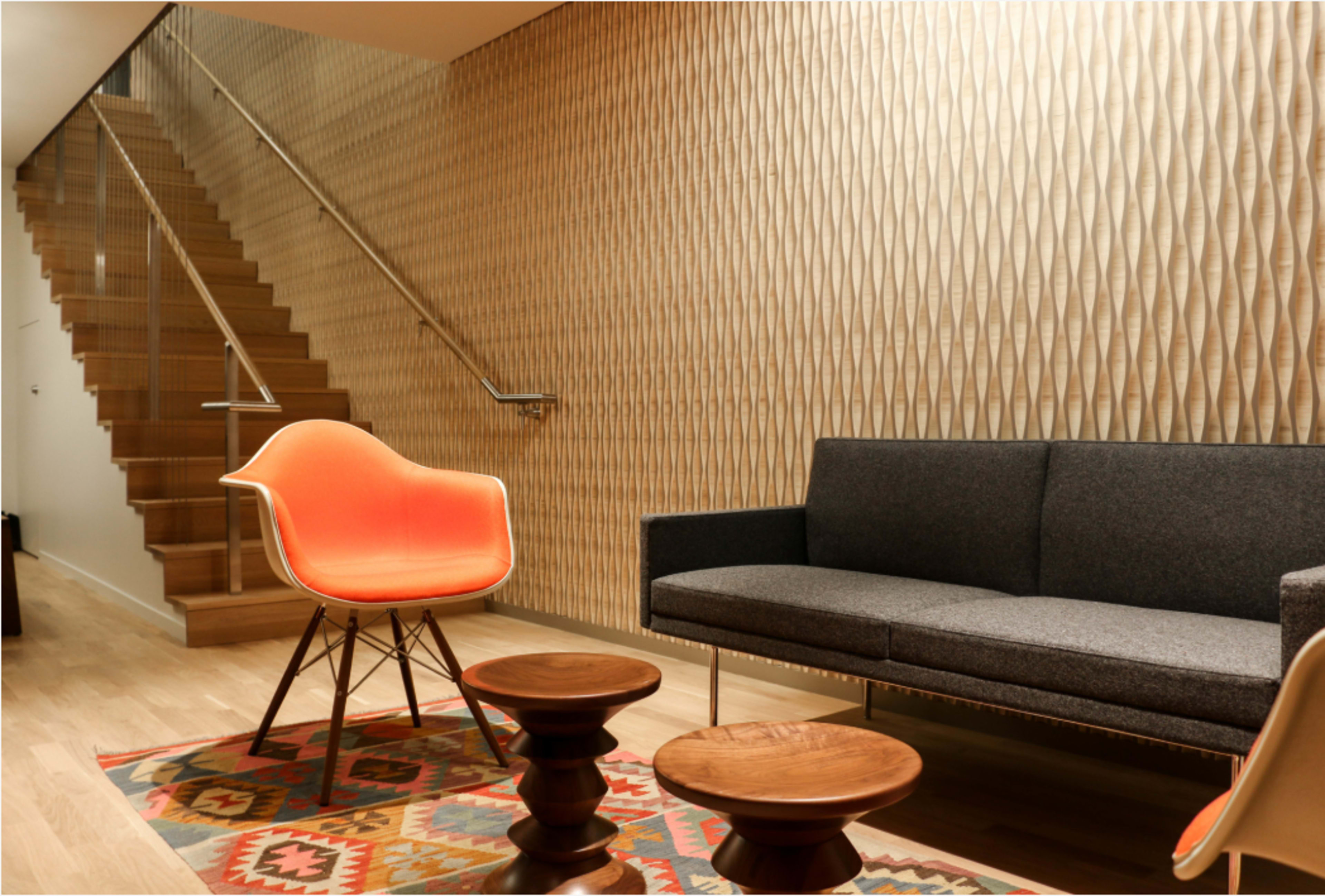 The image shows a modern interior living space with a gray sofa, an orange chair, and wooden coffee tables, alongside a staircase featuring a textured wall.