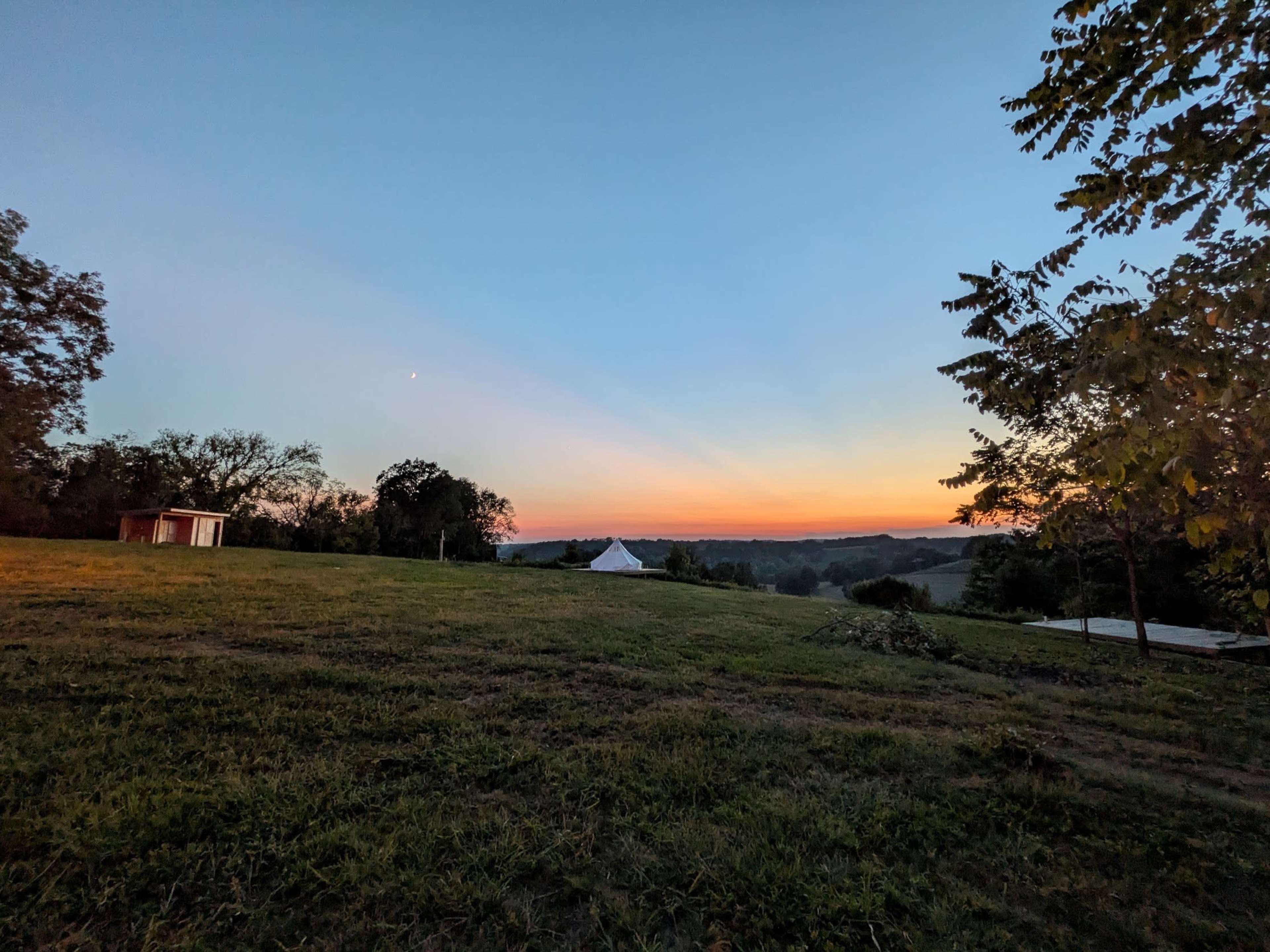 The image shows a grassy landscape at sunset, with a small shed and a tent in the foreground, trees on either side, and rolling hills in the background.