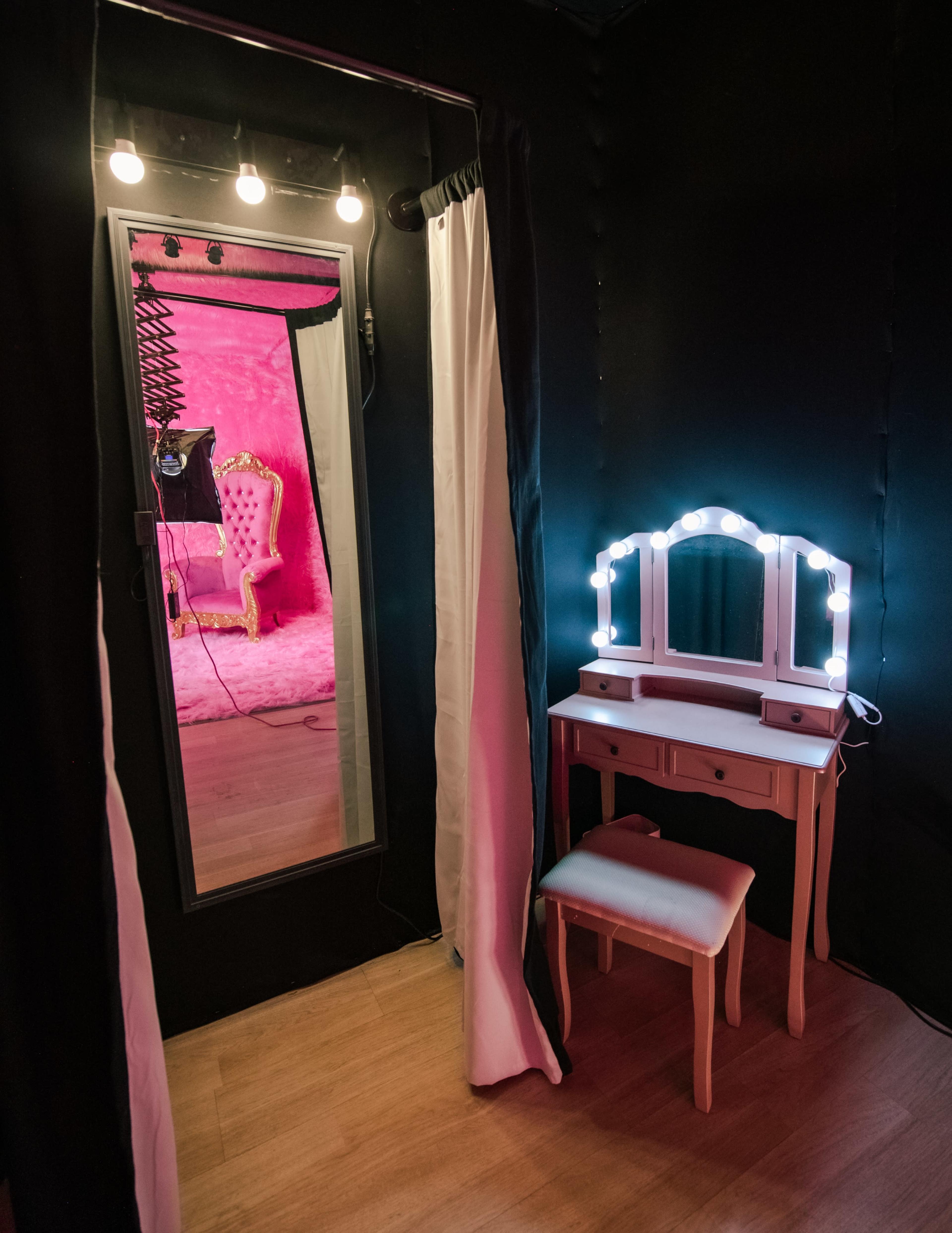 A small vanity with a illuminated mirror sits next to a doorway leading to a brightly colored room with a plush pink backdrop and a decorative chair.