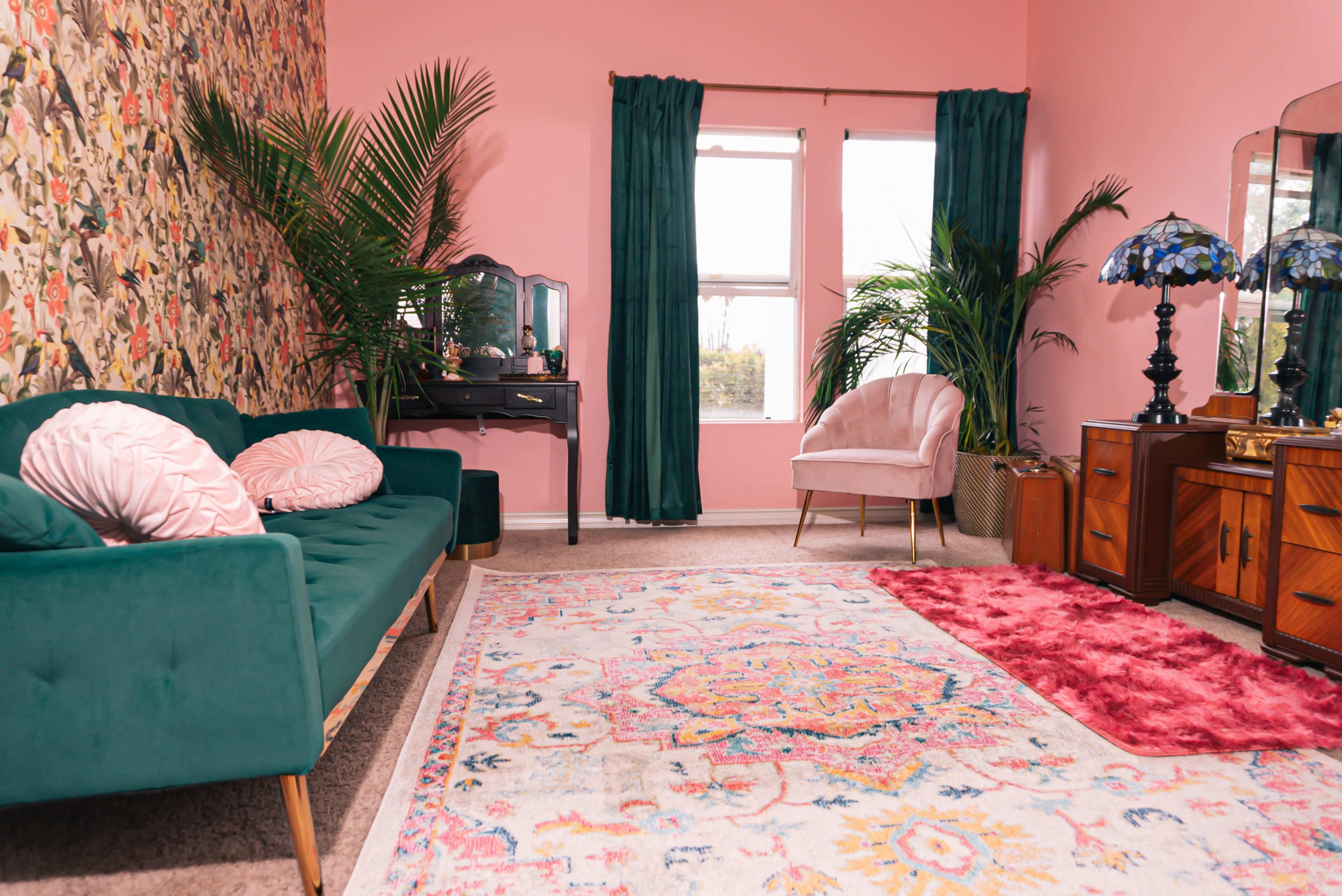 A bright pink room furnished with a green sofa, a light pink chair, a patterned rug, and greenery, complemented by vibrant curtains and a vintage-style dresser.