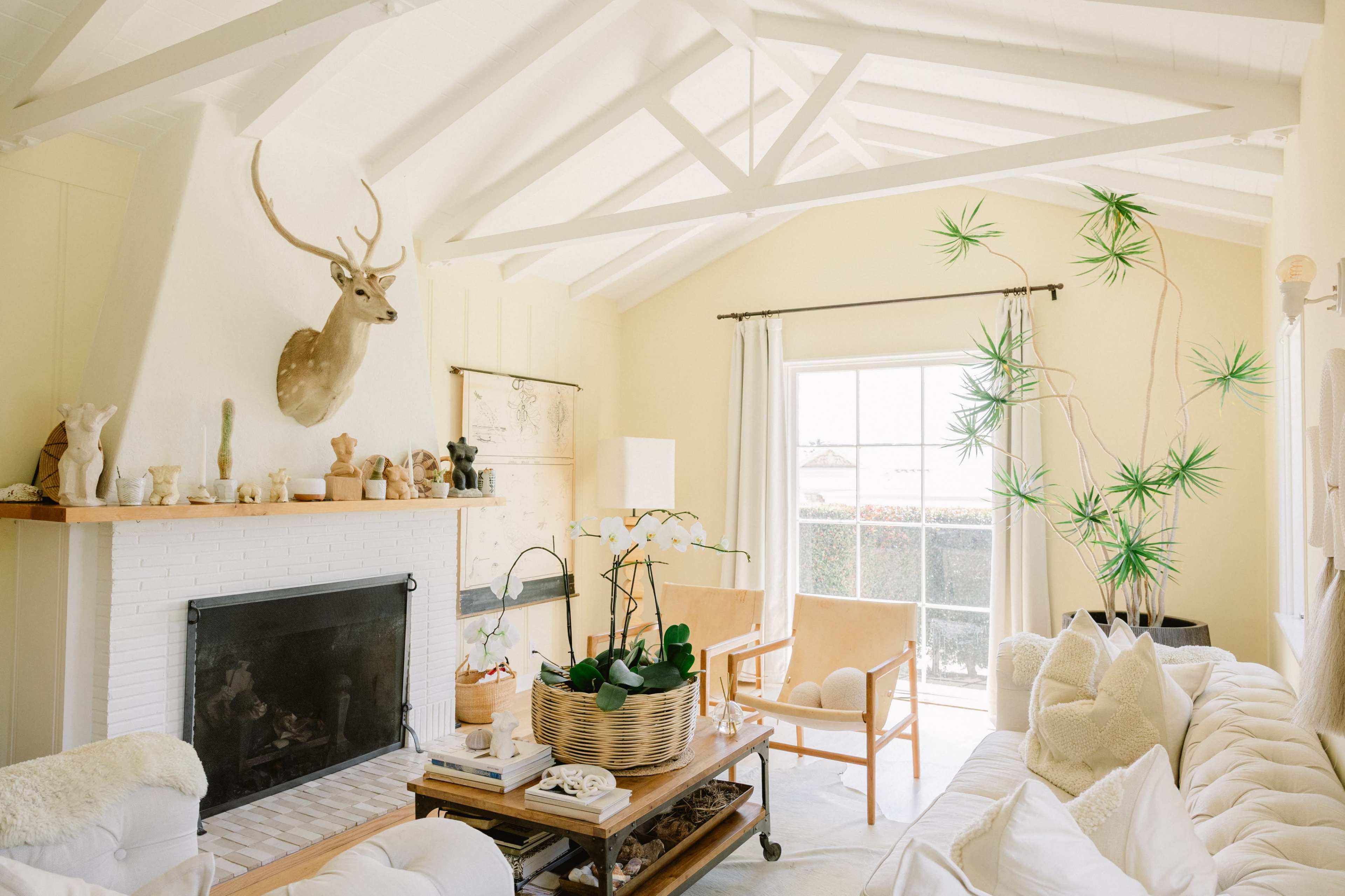 The image shows a bright living room with a high, sloped ceiling, a decorated fireplace, a large window, and a mix of furniture including a sofa and an armchair.