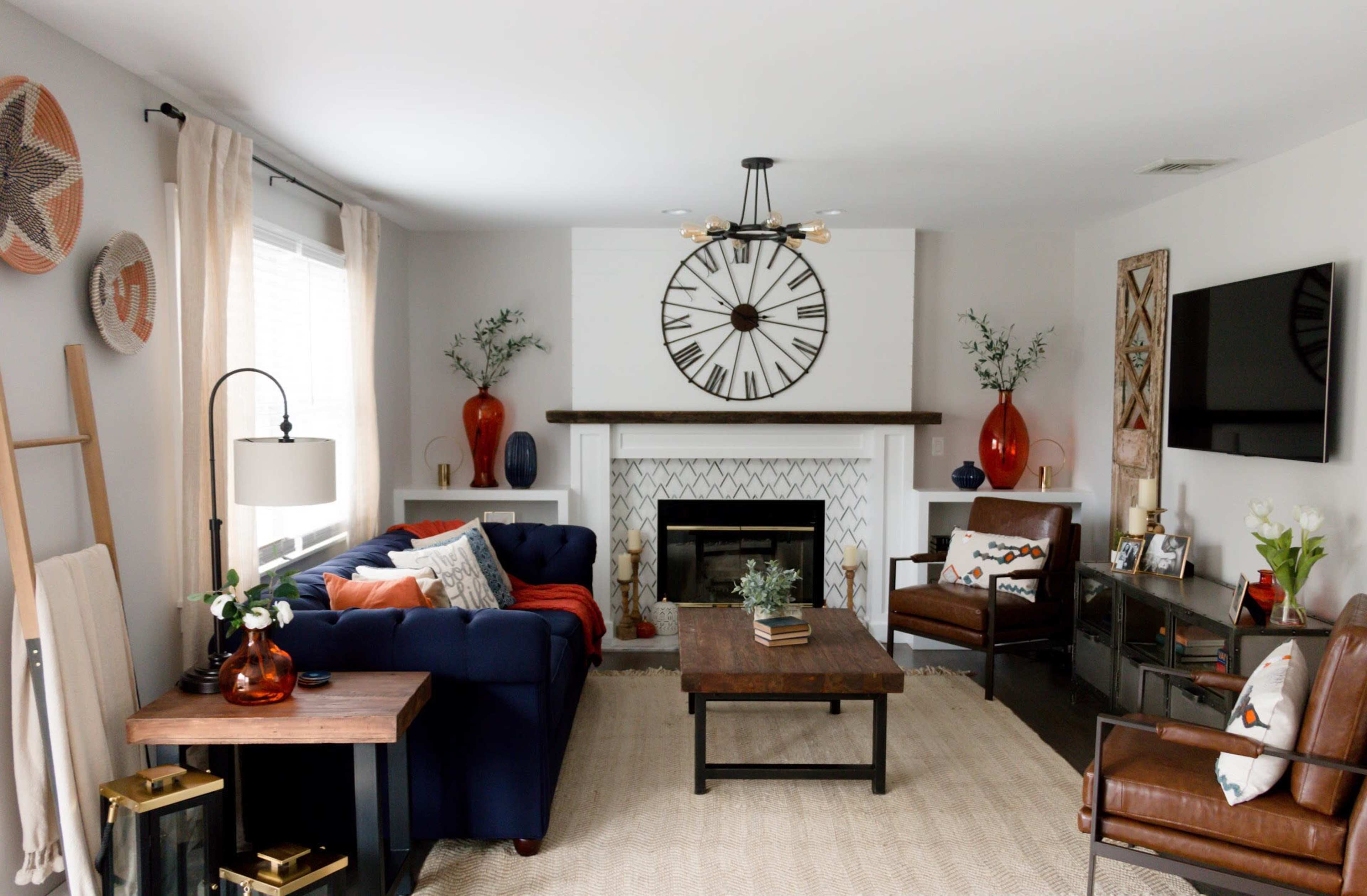 The living room features a blue couch, two brown chairs, a coffee table, and a fireplace with a large clock above it, all arranged around a neutral rug.